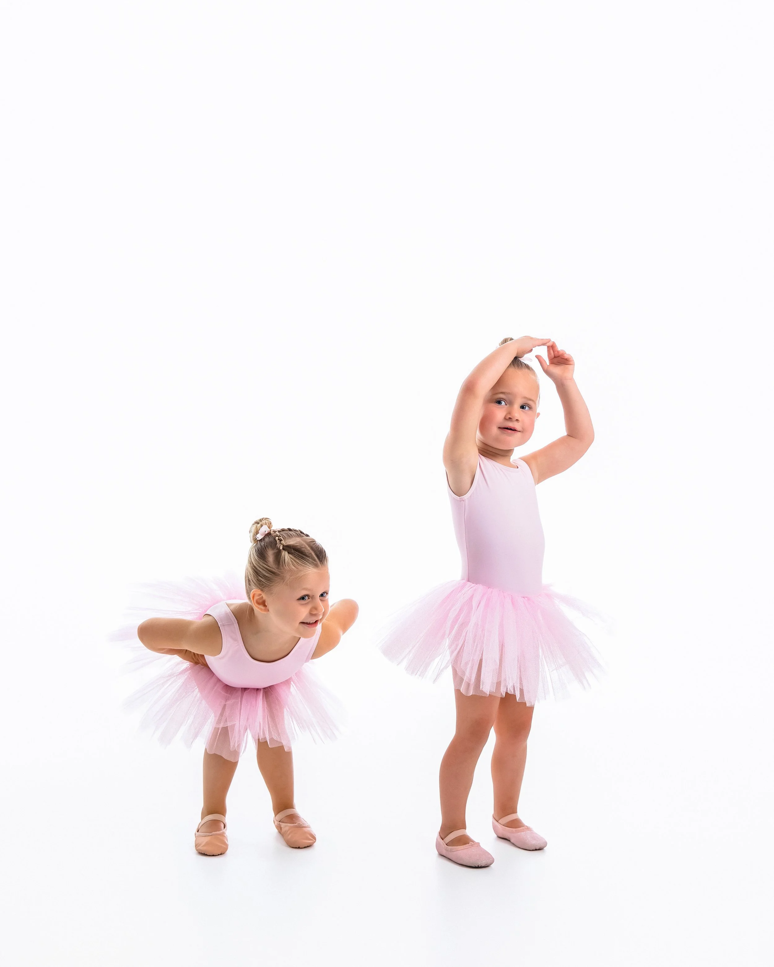 Three young girls wearing pink ballet tutus and shoes, practicing ballet moves against a white background.