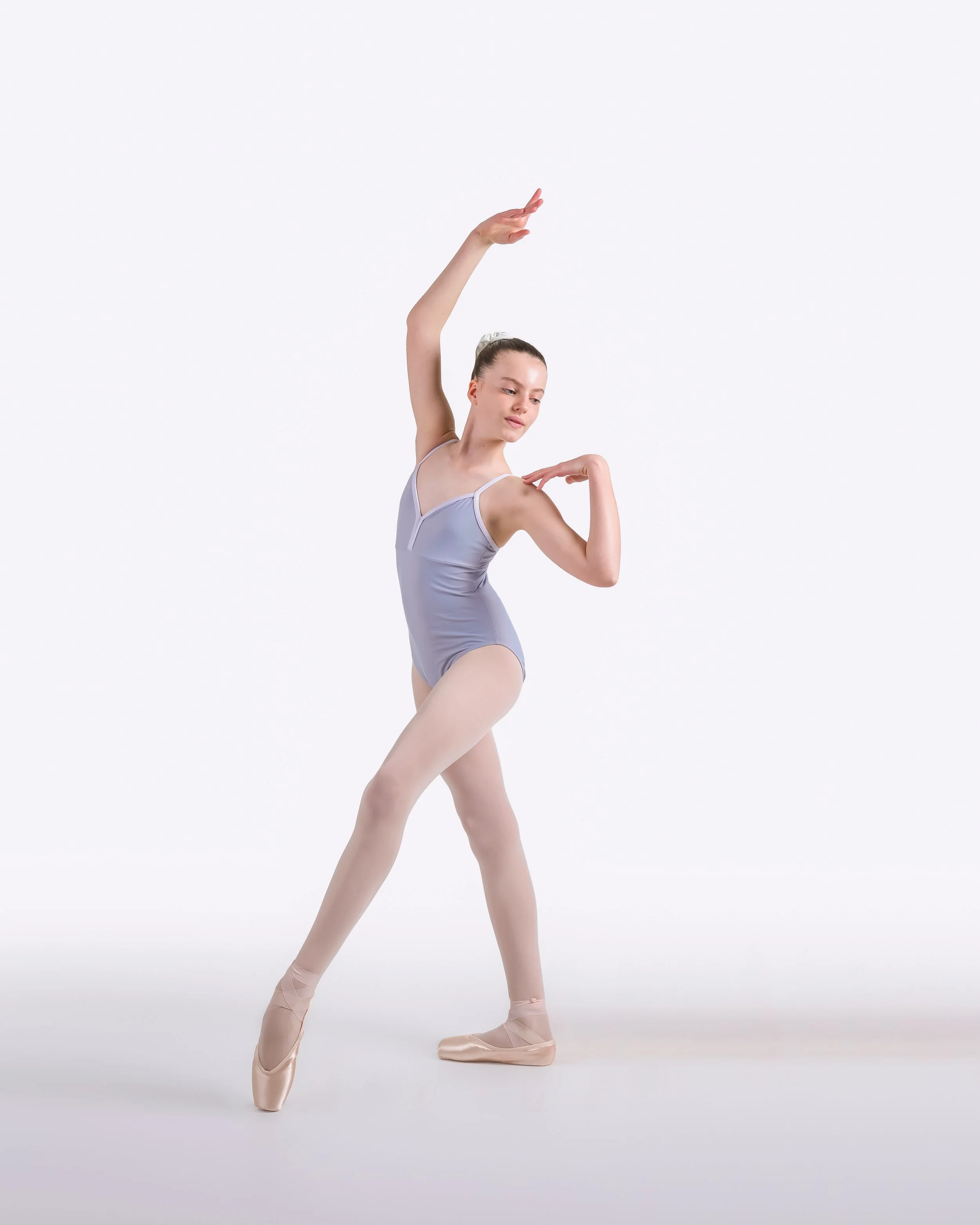A ballerina dressed in a grey leotard and pink pointe shoes performs a dance pose against a plain white background, with one arm raised above her head and the other bent at the elbow near her shoulder.
