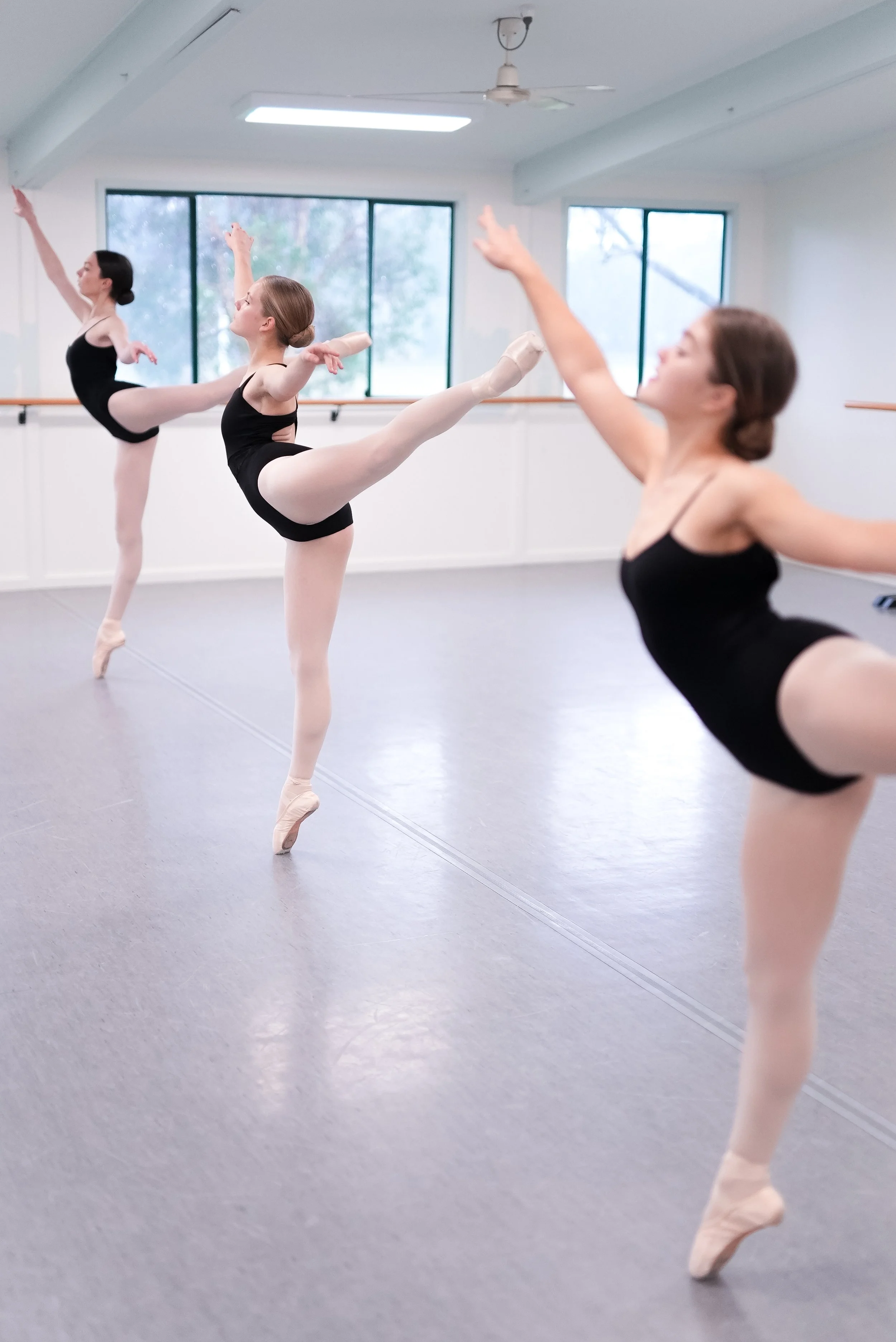 Three ballet dancers practicing in a dance studio, wearing black leotards and pink ballet slippers, performing a high kick with arms raised.