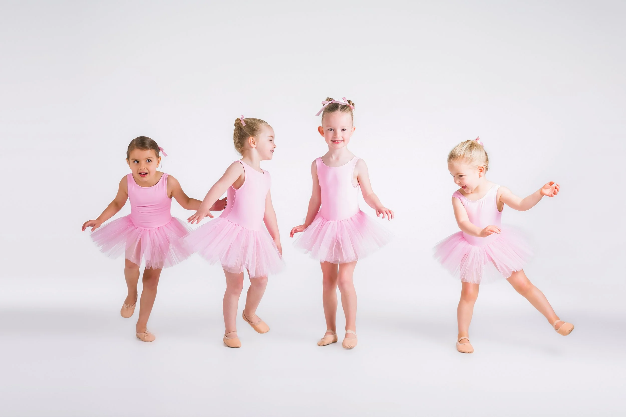 Five young girls dressed as ballerinas in pink tutus and pink bows, dancing against a white background.