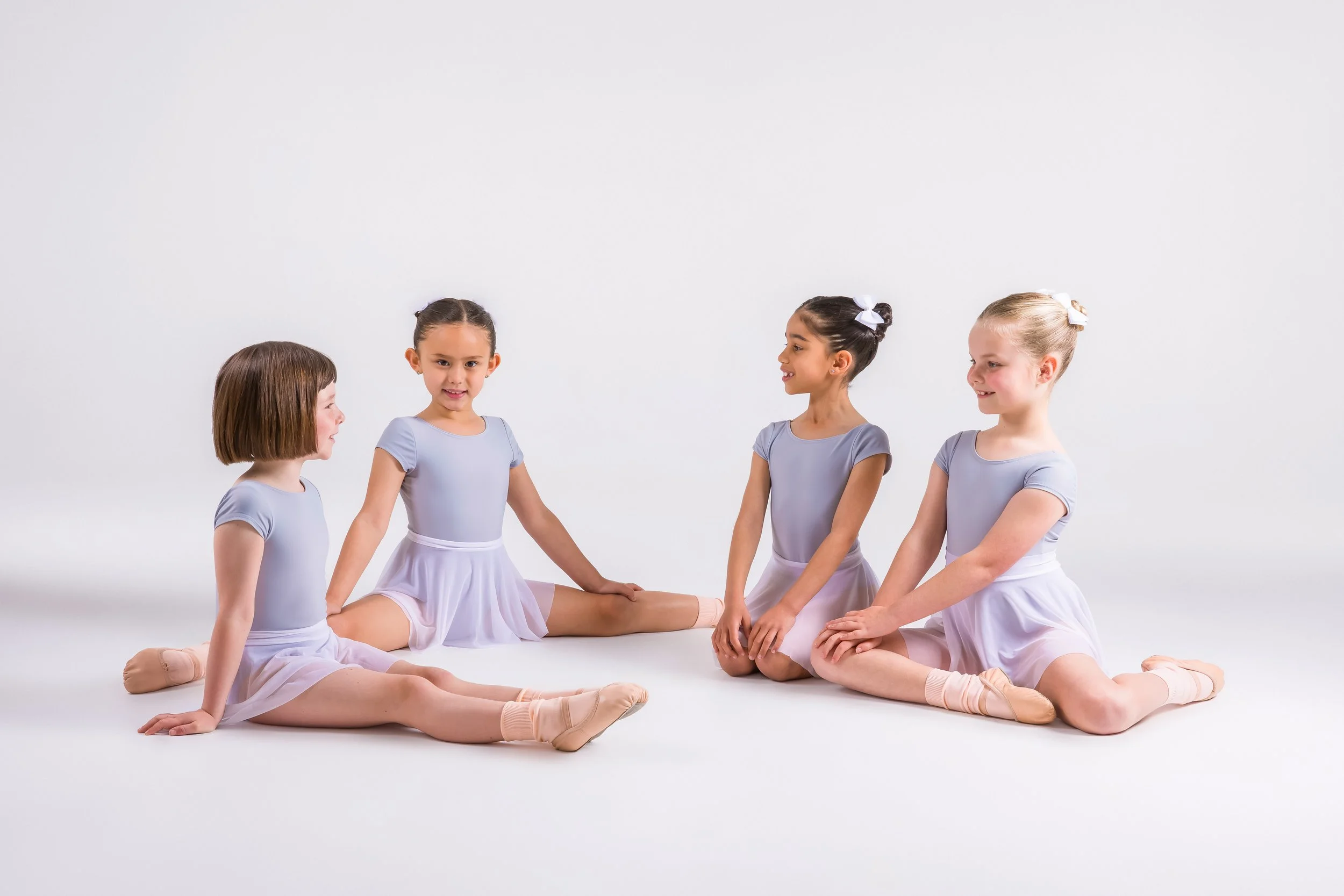 Four young girls dressed in ballet outfits sitting on the floor and engaging in conversation.