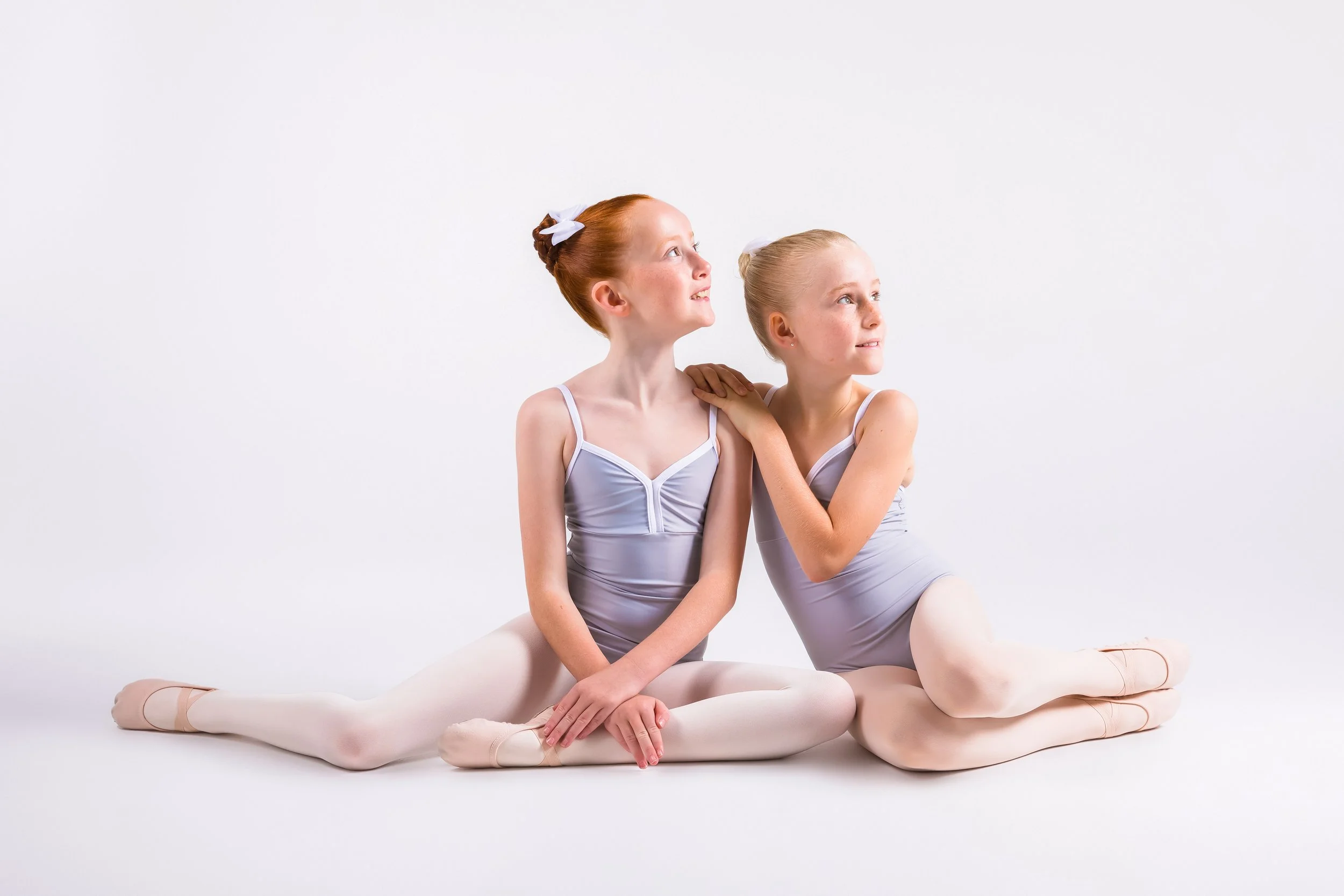 Two young ballet dancers, one with red hair and the other with blonde hair, sitting close together on the floor in ballet practice wear, looking up and to the side, in front of a plain white background.