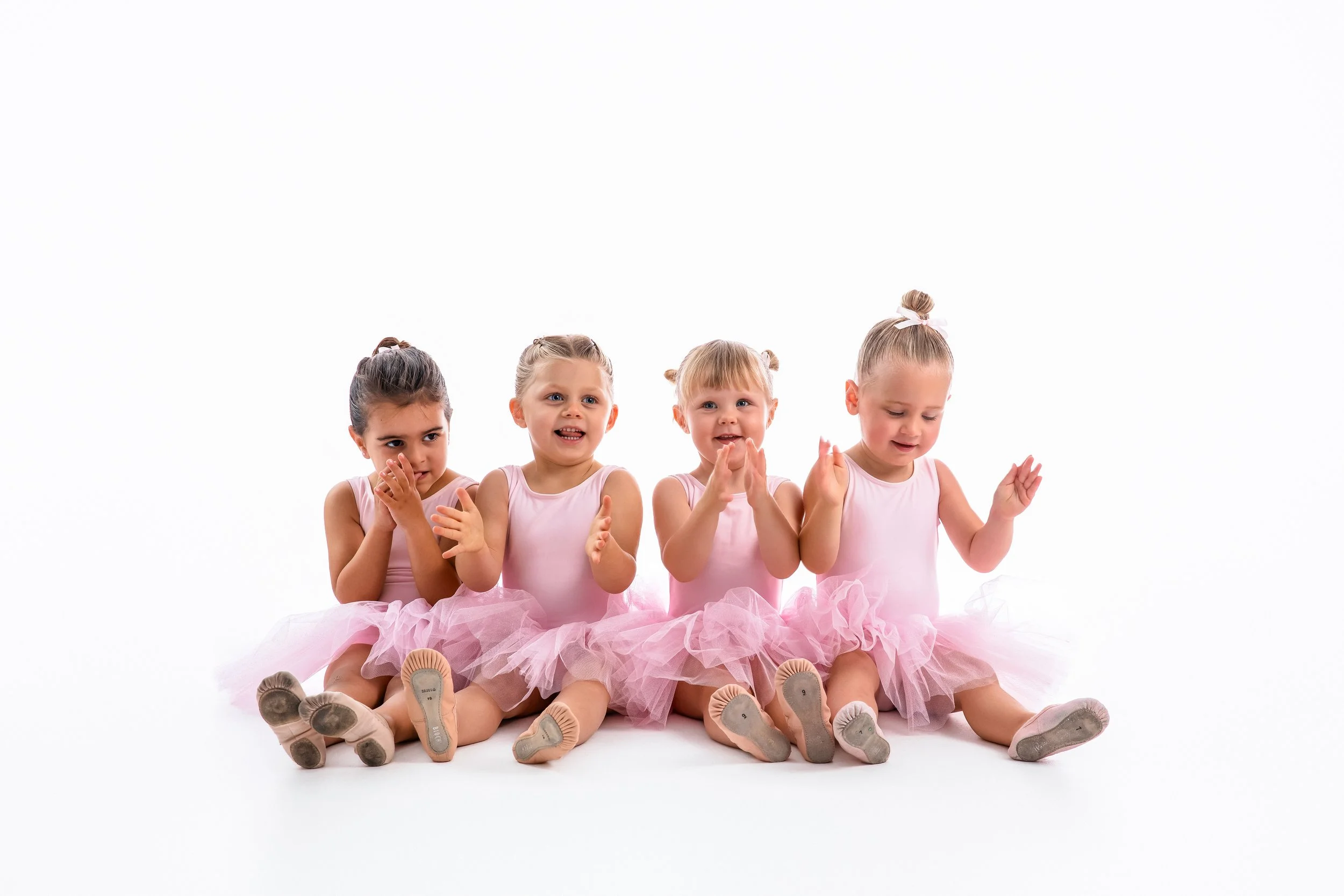 Four young girls dressed as ballerinas sitting on the floor, clapping and smiling, with a plain white background.