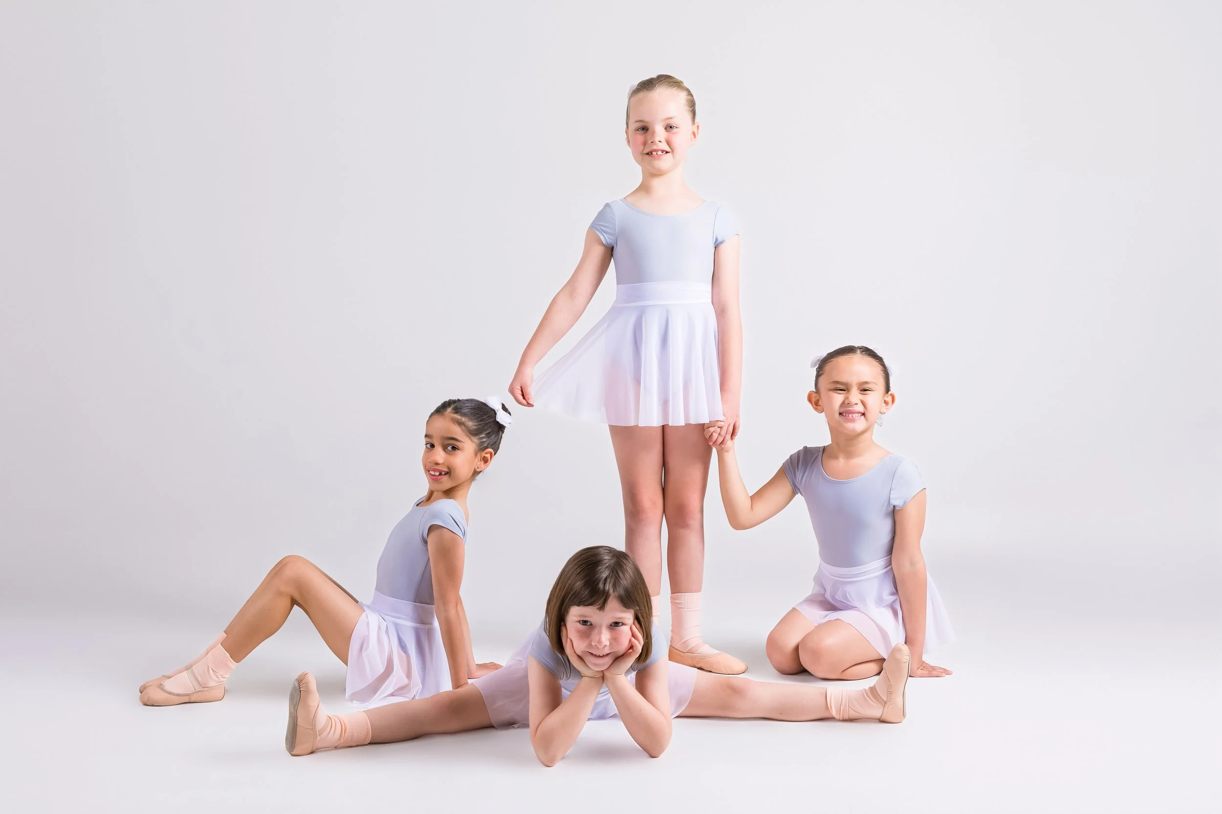 Group of five young girls in dance costumes posing in a ballet studio against a plain light gray background.
