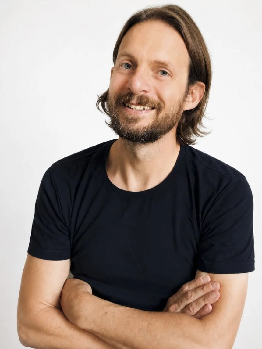 Ein lächelnder Mann mit braunen Haaren, Bart und blauen Augen, trägt ein schwarzes T-Shirt, steht vor einem weißen Hintergrund.