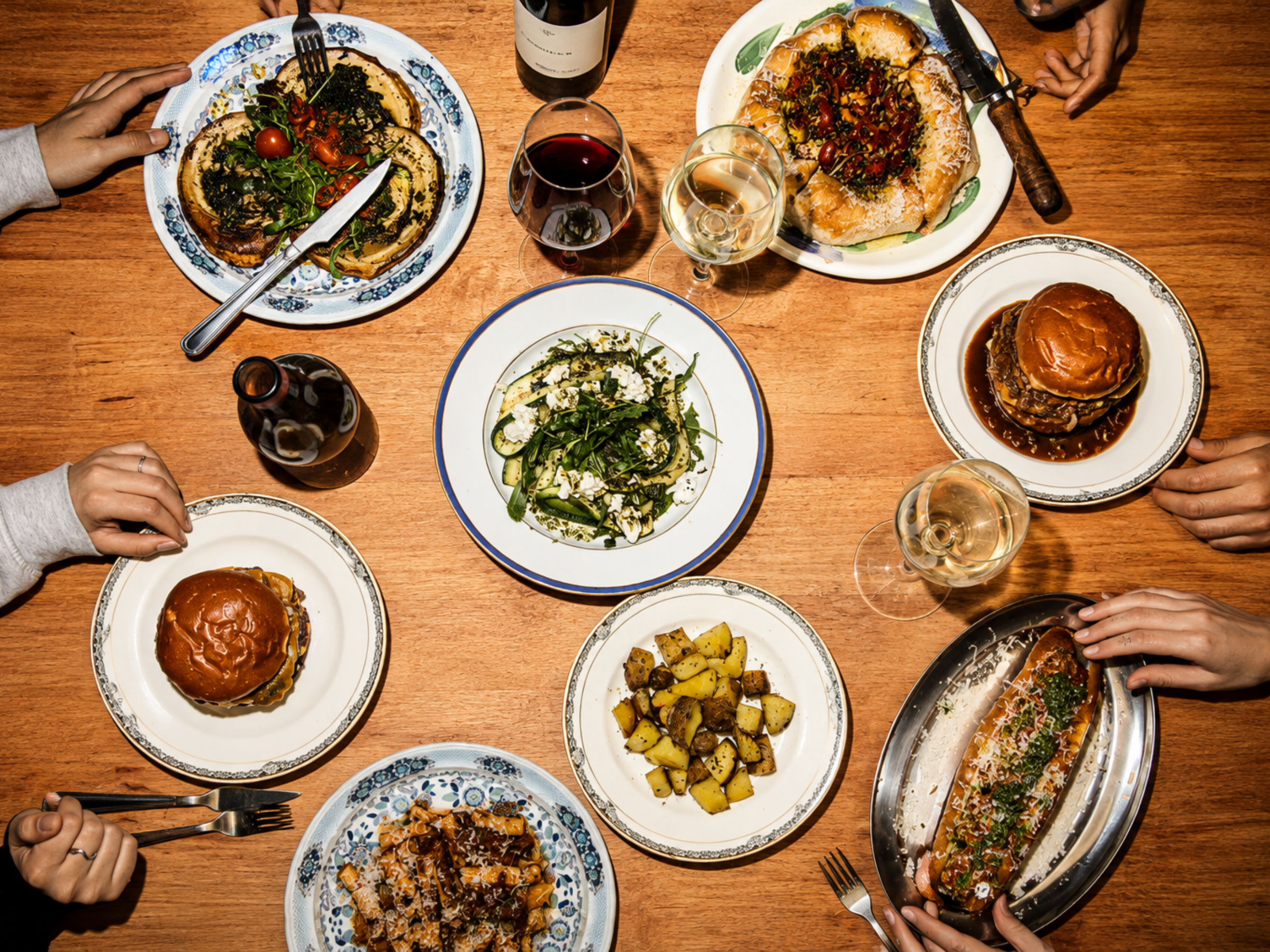 A variety of dishes on a wooden table, including a burger with tomato and greens, a salad with lemon dressing, roasted potatoes, a pizza, and a long tray with a rolled wrap and garnishes, along with glasses of red and white wine.