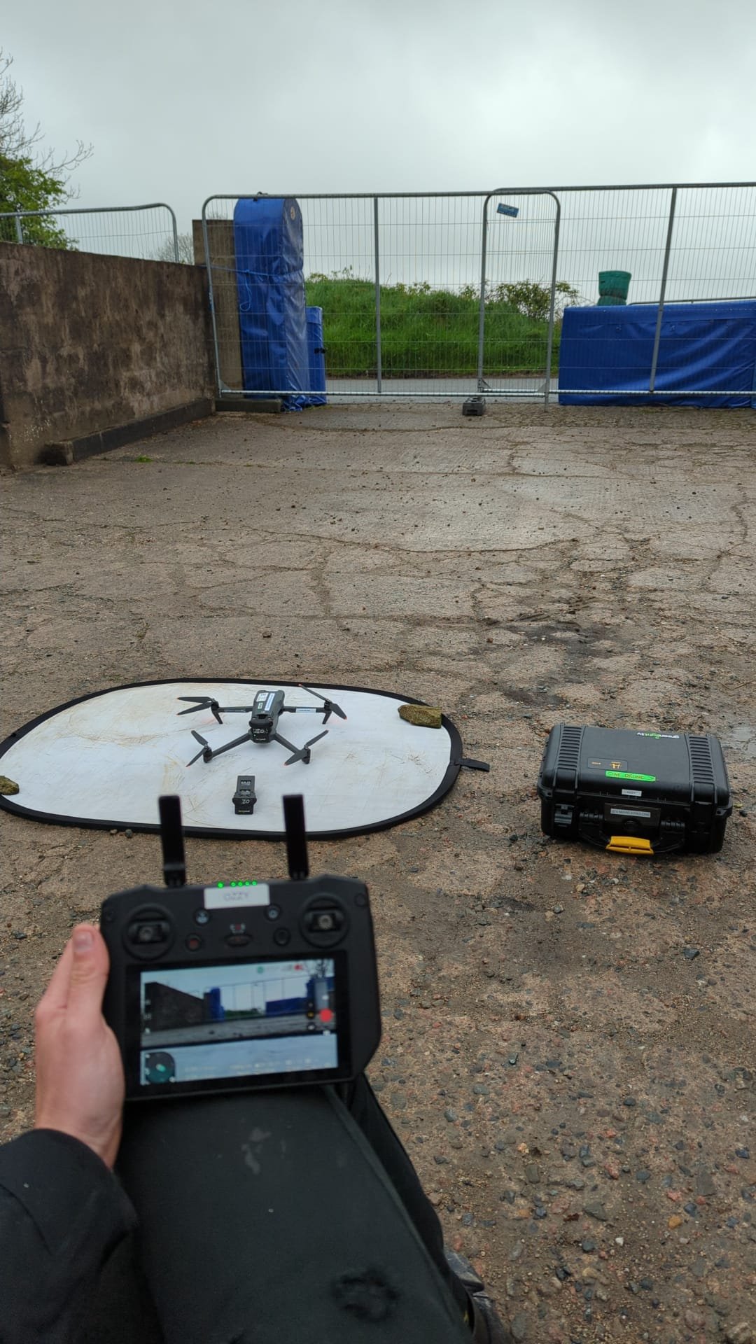 A drone on a white landing pad with a remote control in the foreground, outdoors on a cracked pavement with a fenced area and green grass in the background.