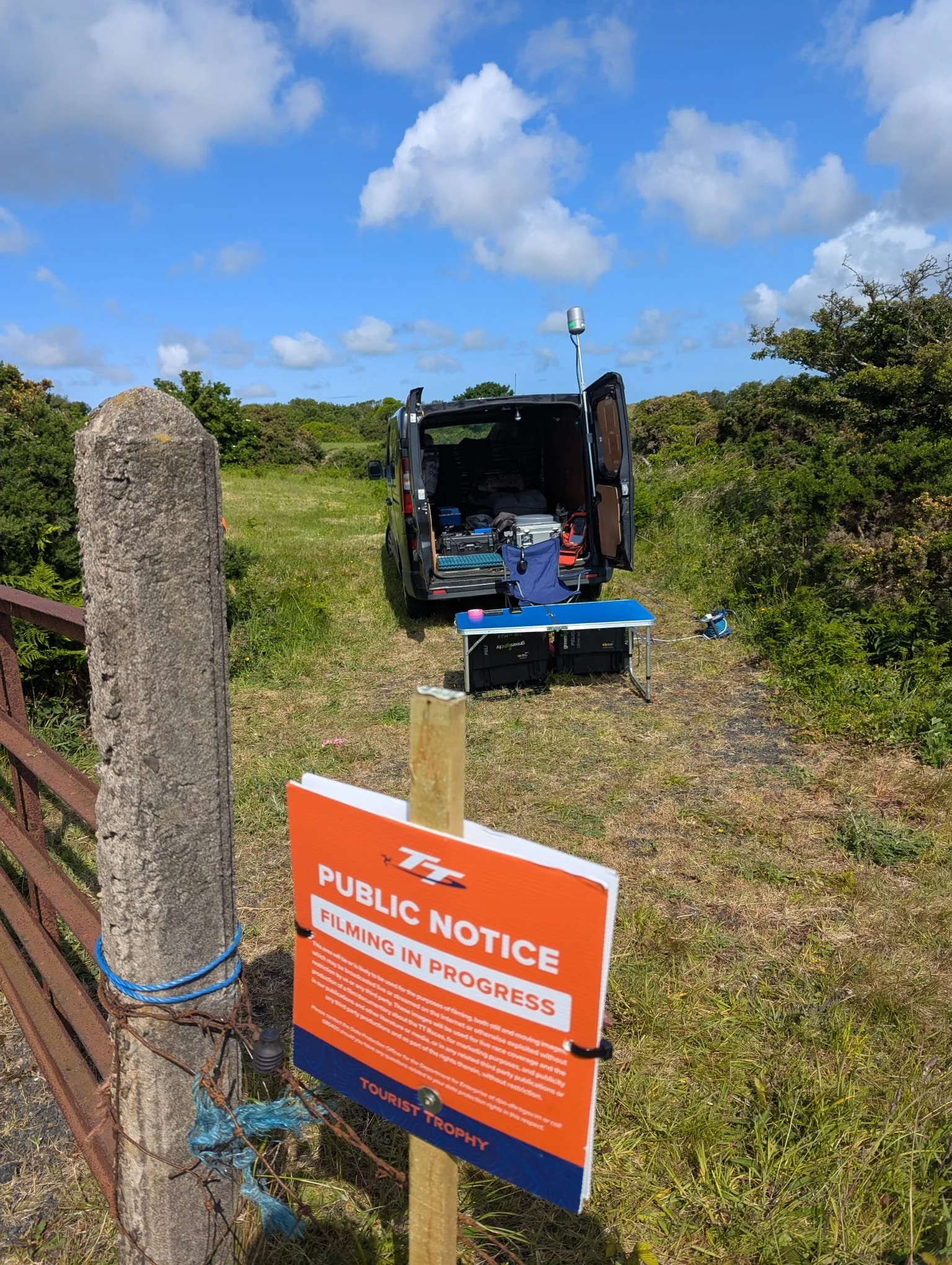 A black van with its back doors open parked on a grassy area under a partly cloudy sky. Equipment and supplies are visible inside the van. In the foreground, there is an orange and blue notice sign attached to a wooden post and a weathered stone pole wrapped with blue twine.