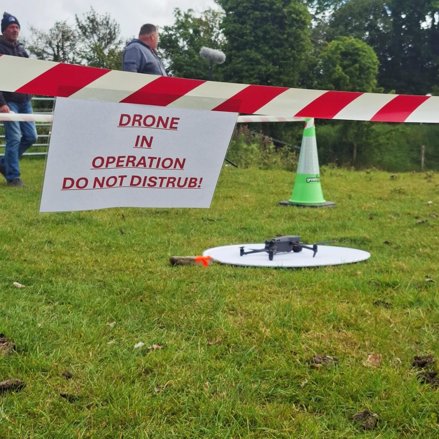 A drone on the ground in a field, with a sign warning 'Drone in Operation Do Not Disrupt,' a green traffic cone, and a group of people in the background.