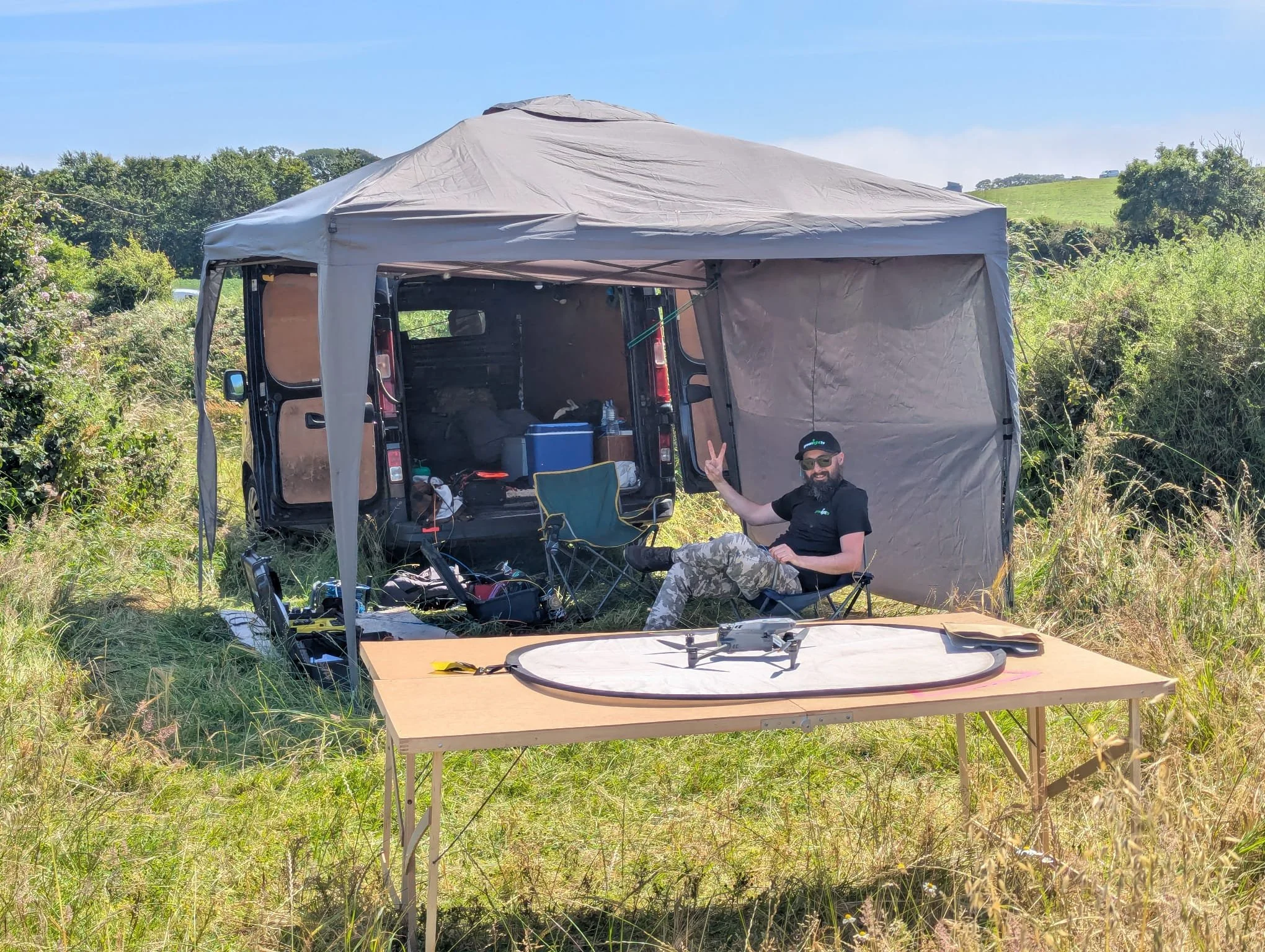 A man with a beard, sunglasses, black T-shirt, and camouflage pants sitting in a camping chair outdoors near a van with a canopy, surrounded by tall grass and trees, under a clear blue sky.