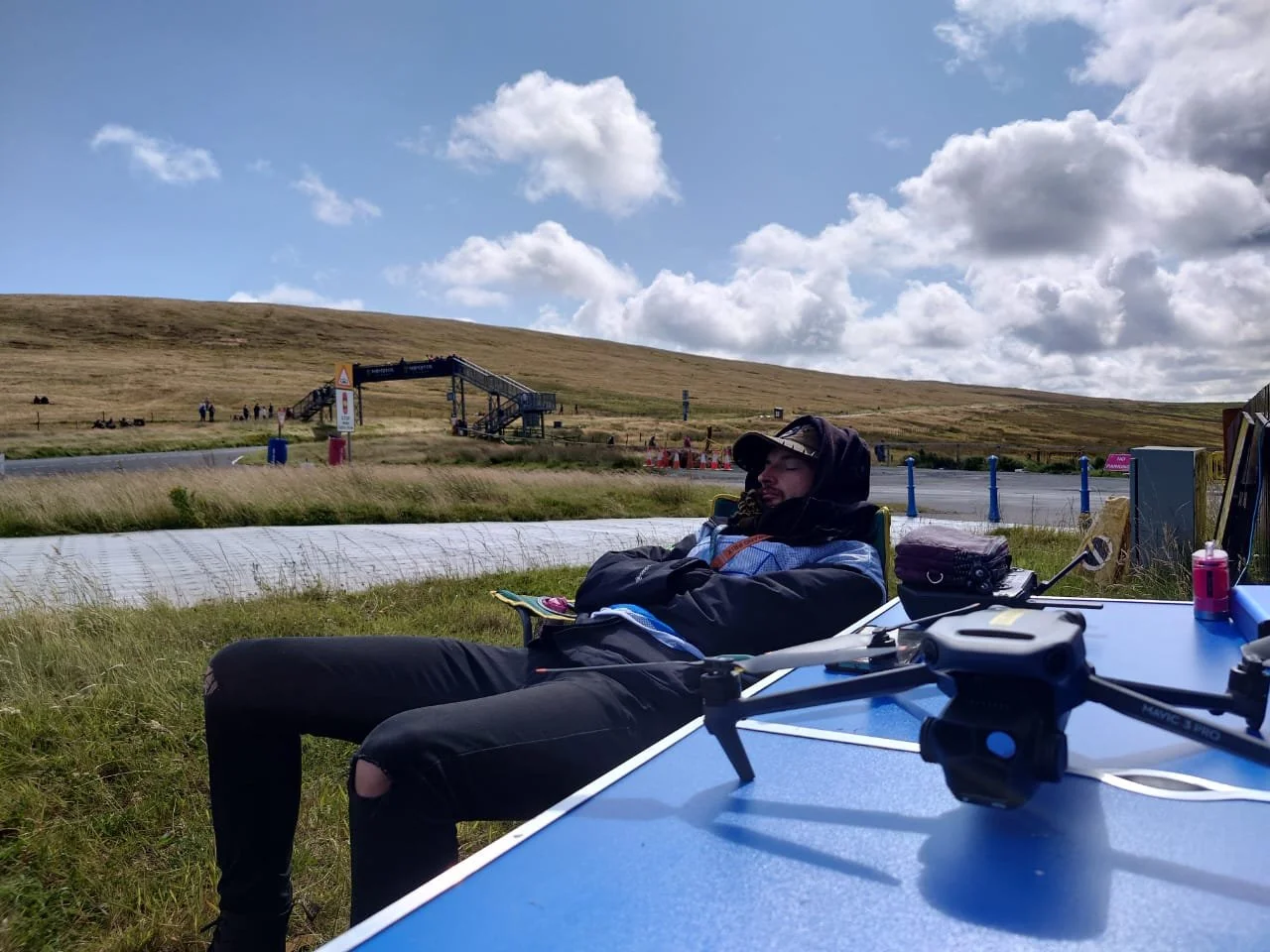 Person sitting outdoors under a cloudy sky with a drone on a table in front. They are wearing a hoodie and sunglasses, relaxing on a chair with a backpack nearby. In the background, a grassy hill and a walkway with some people and a staircase can be seen.