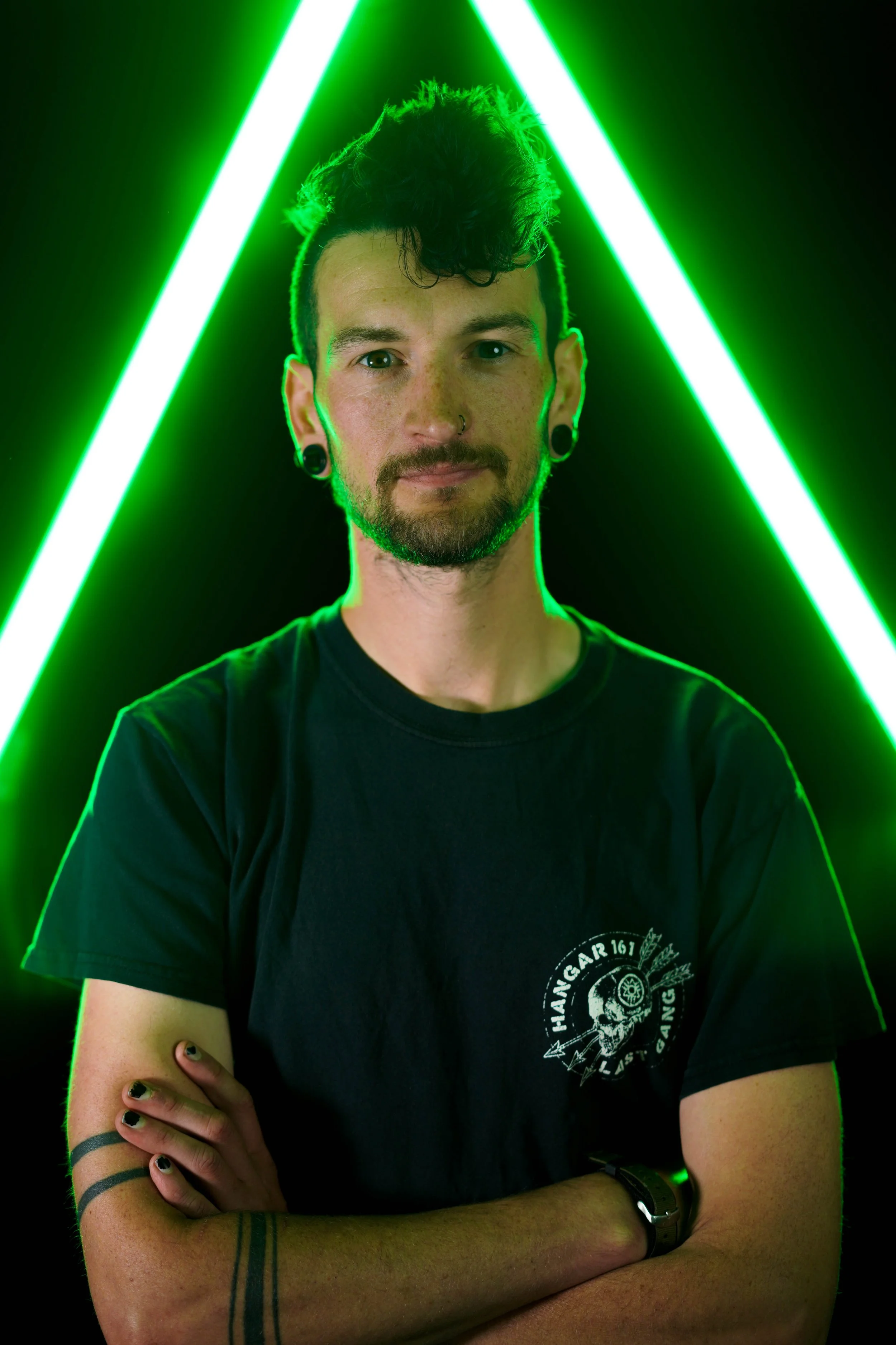 A young man with short dark hair styled into a mohawk, sporting piercings and tattoos, wearing a black t-shirt with a skull design. He is standing with his arms crossed against a black background illuminated by vibrant green neon lights forming an 'A' shape.