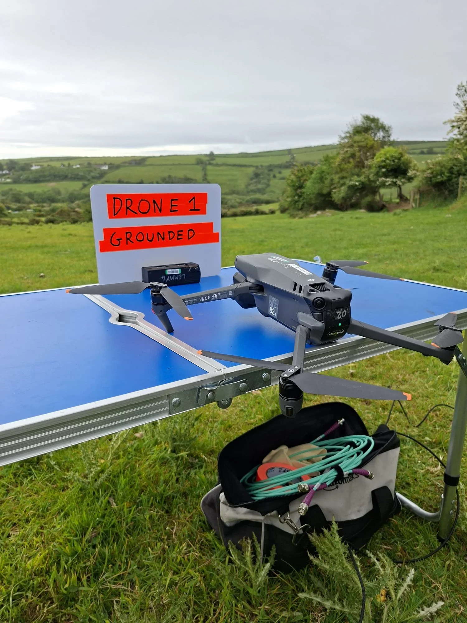 A drone with landing gear resting on a table outdoors, with a sign reading 'DRONE 1 GROUNDED' in the background, situated in a grassy open field with rolling hills and trees under a cloudy sky.