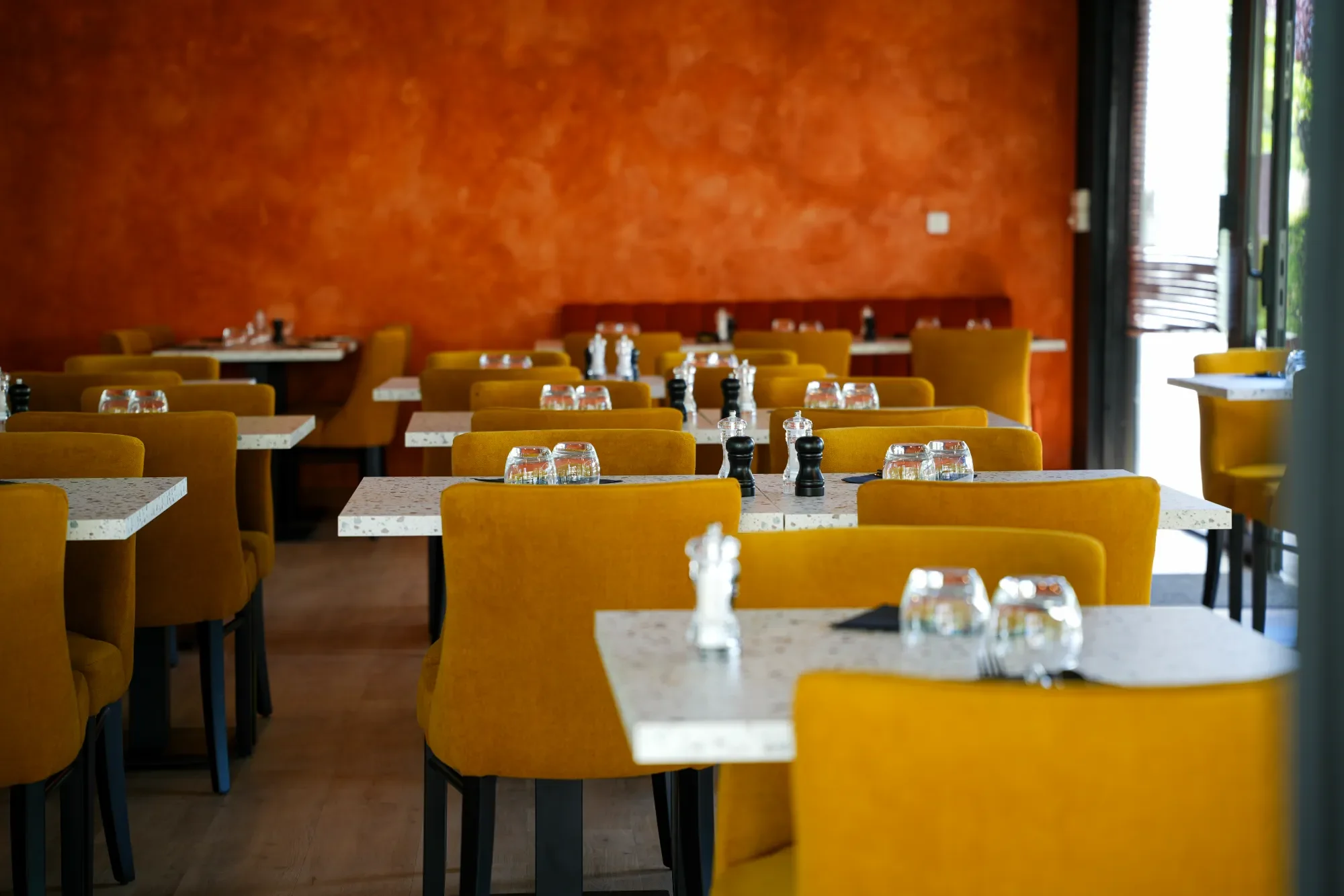 Salle à manger de restaurant avec murs orange, tables en marbre blanc, chaises jaunes, et assiettes, verres, poivriers, et sel sur les tables, près de grandes fenêtres laissant entrer la lumière.