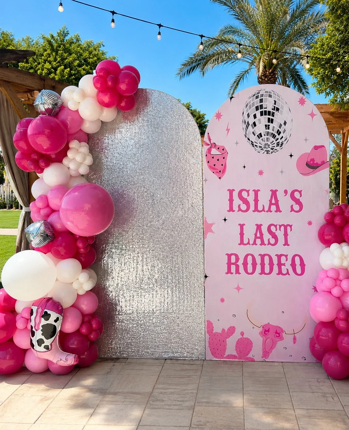 Decorative backdrop with pink and white balloons, a silver textured panel, and a pink sign that reads "Isla's Last Rodeo" with cowboy-themed illustrations, set outdoors with palm trees and string lights overhead.