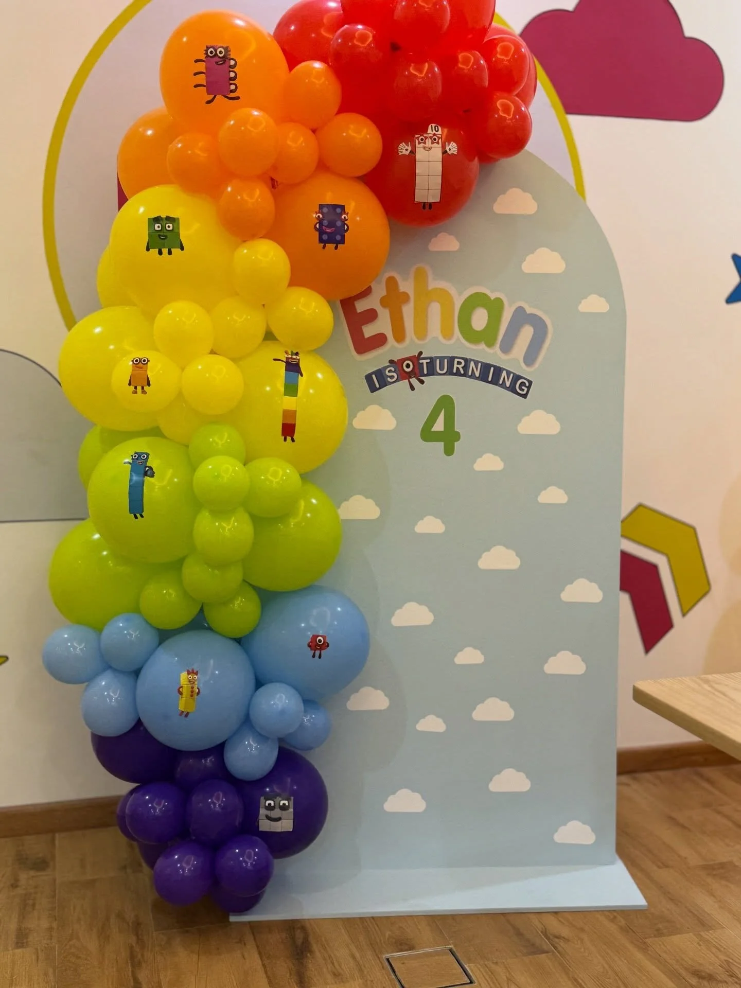Colorful balloon display arranged in rainbow colors from purple to red, with cartoon monster stickers, on a backdrop with clouds and birthday message for Ethan's 4th birthday.