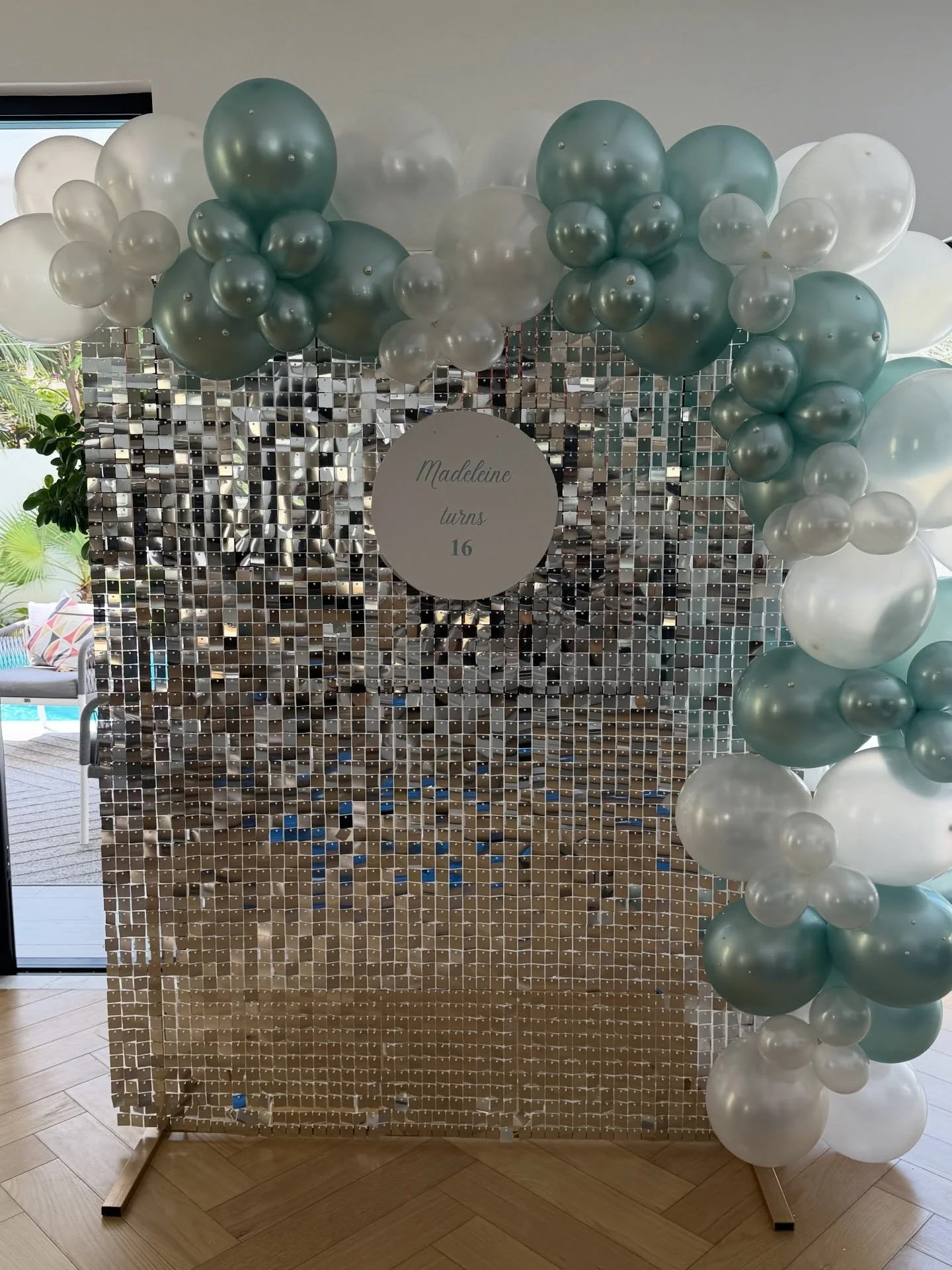 Decorative backdrop with a silver mosaic tile wall, a balloon arch with white and teal balloons, and a round sign that reads "Madeleine turns 16" in the center.
