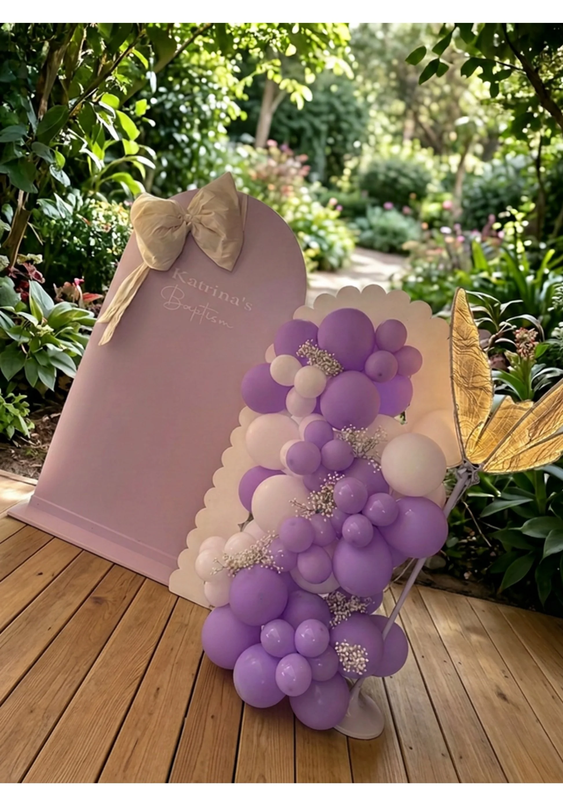A pastel purple woode backdrop with a cream bow decorated with purple and white balloons and flowers, set outdoors perfect for winter gatherings