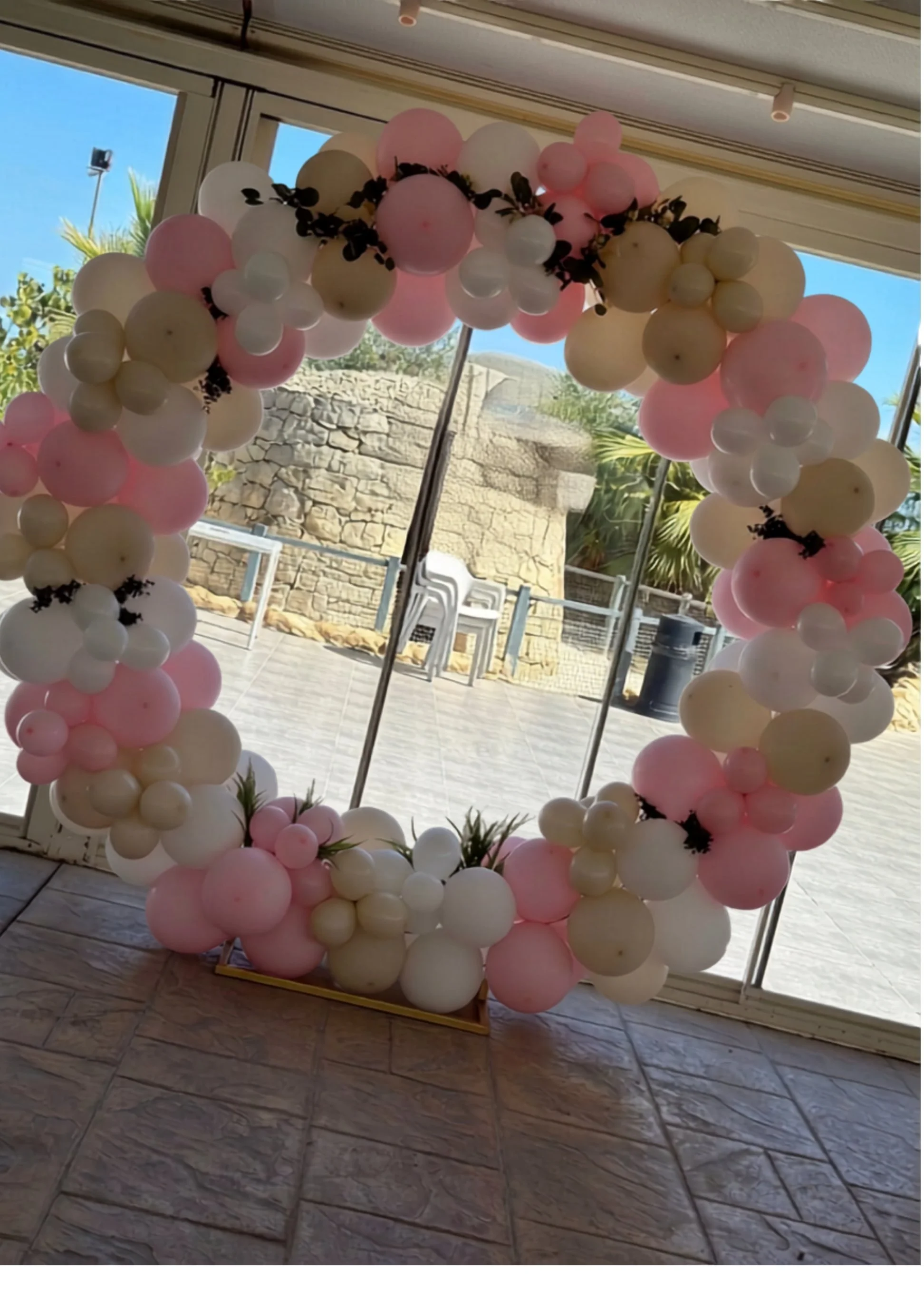 Balloon arch in white, pink, and beige colors decorated with black leaves, set up at an indoor event space with large windows showing outdoor scenery of blue sky, trees, and stone wall.