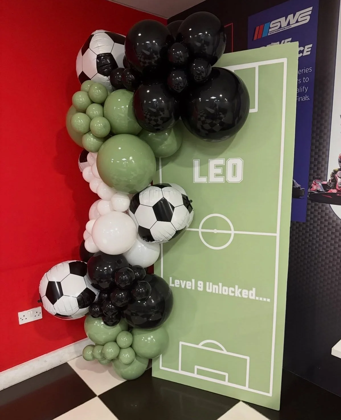 A football ground backdrop of black, white, and green balloons arranged with football shaped balloons for a footy fanatic.