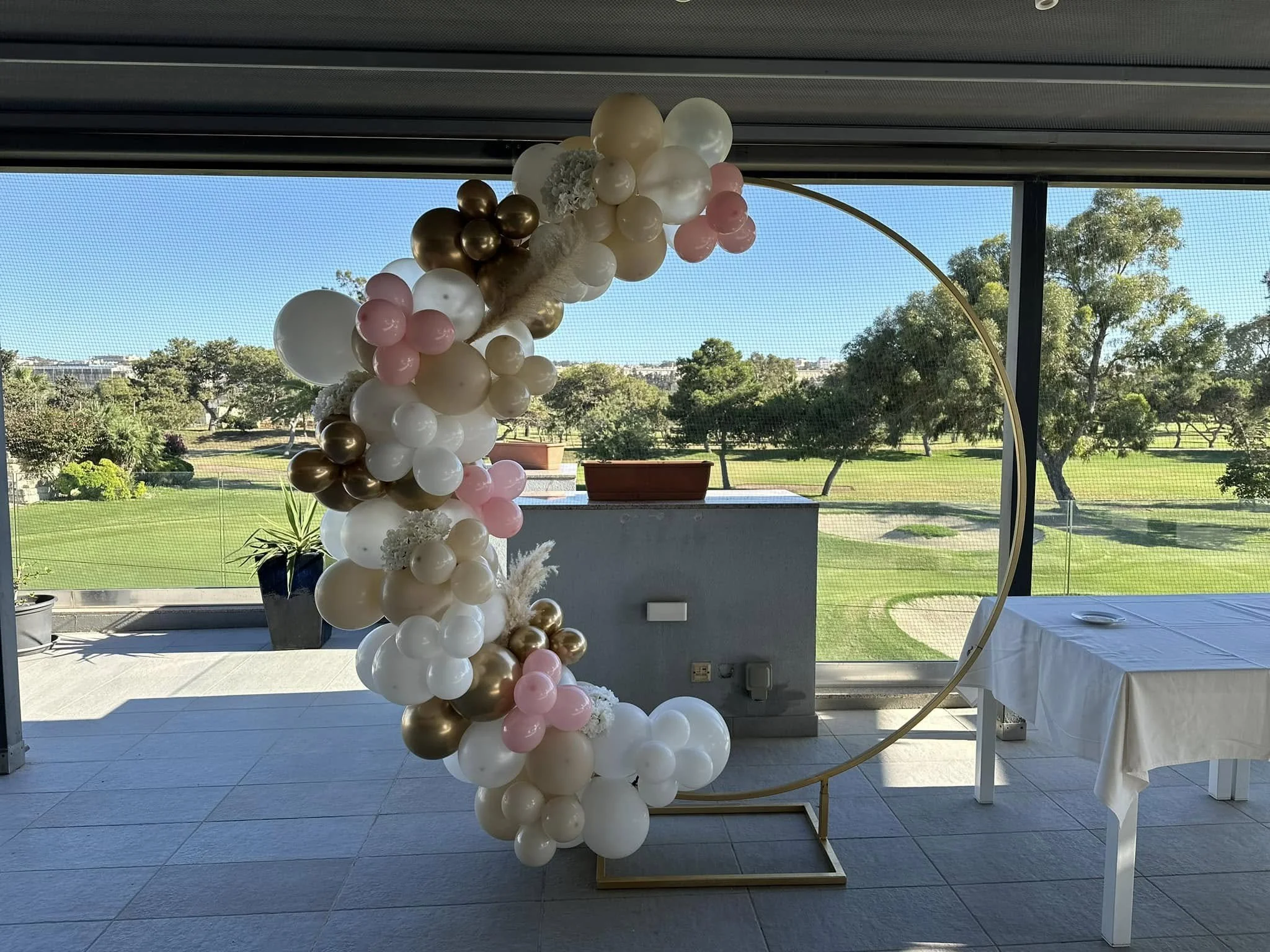 Balloon arrangement with white, pink, gold, and pearl-colored balloons, creating an arch, positioned by a window with a outdoor park scene.