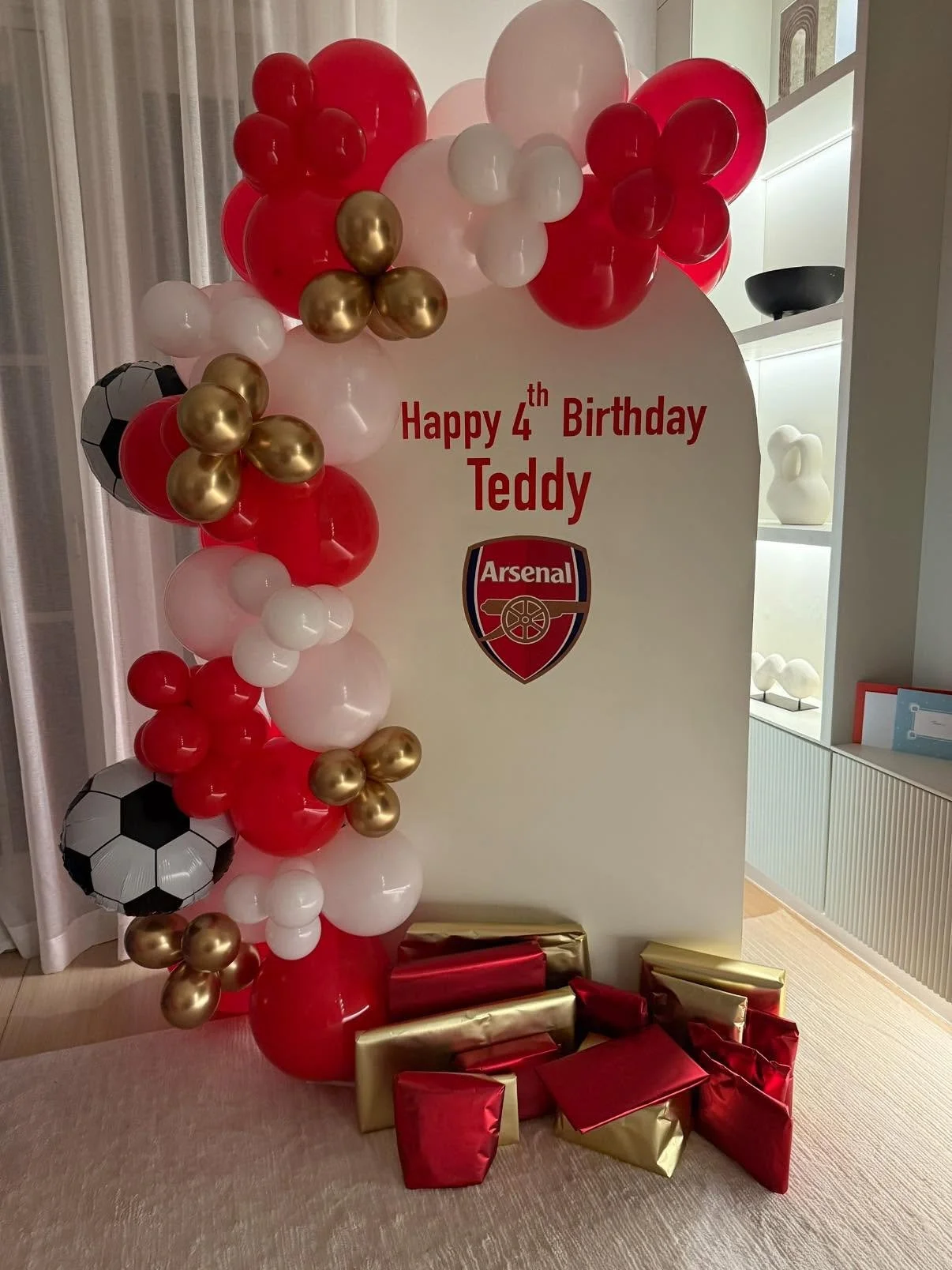 Birthday display with balloons in red, white, and gold, featuring Arsenal football club logos, a sign reading "Happy 4th Birthday Teddy," and wrapped gifts at the base.