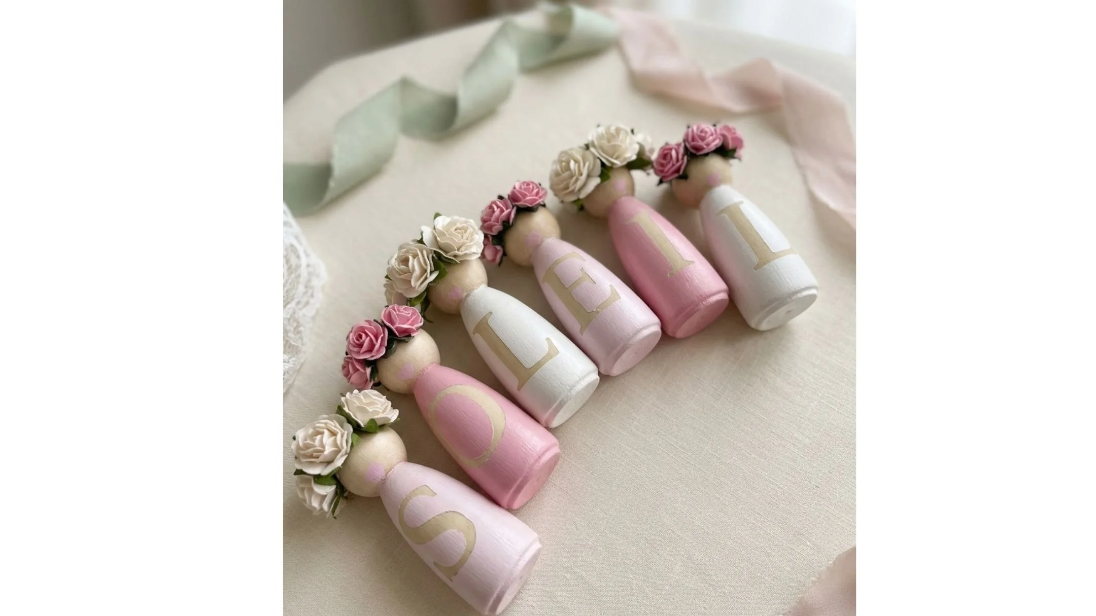 Decorative wooden dolls in pink and white colors, adorned with small floral crowns made of roses.