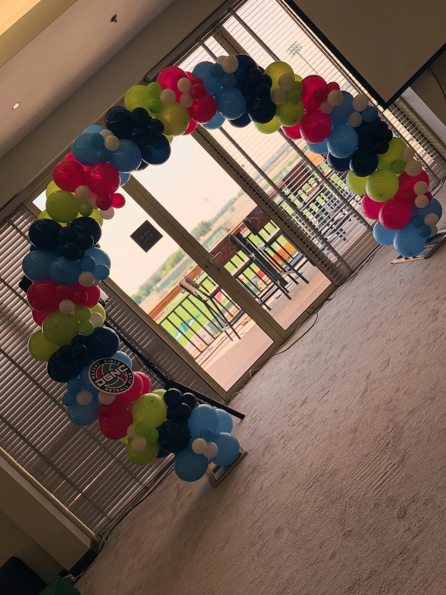 Colorful balloon arch with various colors, including blue, pink, black, green, and white, set up indoors near glass doors with window blinds.