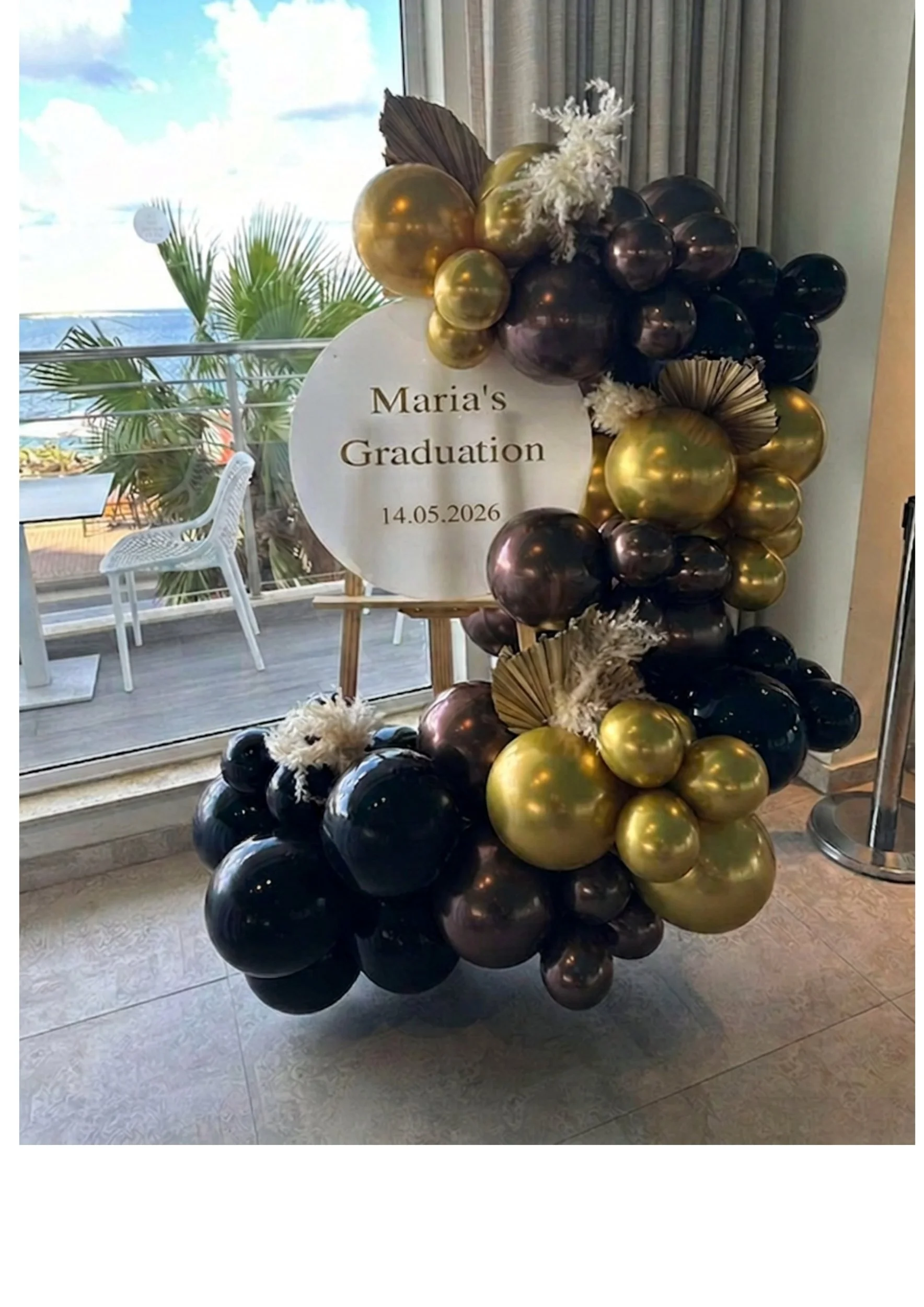 Decorative balloon display with black, gold, and purple balloons, a round sign reading 'Maria's Graduation 14.05.2026', and white flowers, located indoors near a large window with a view of palm trees outside.