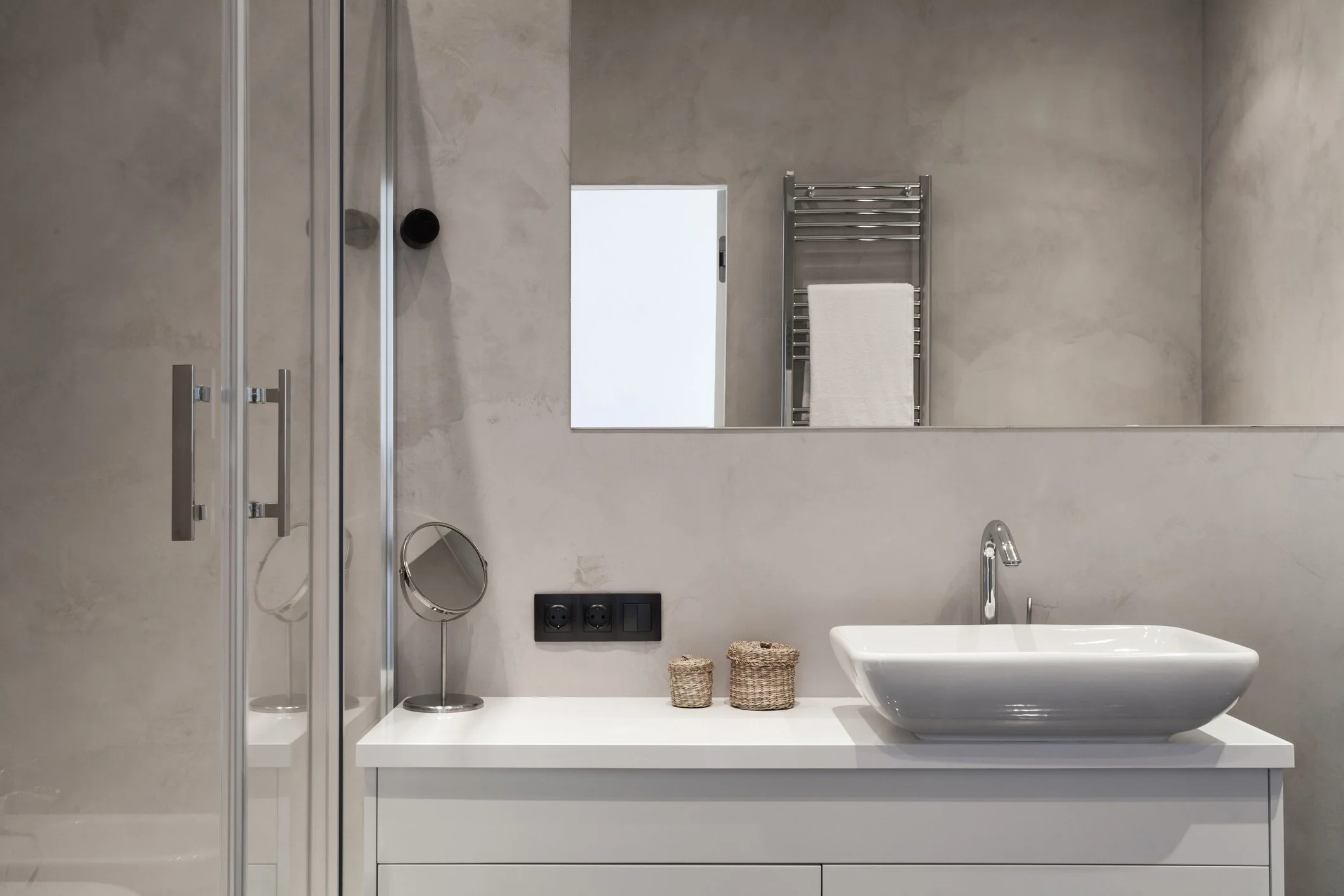 Moderner Badezimmerblick mit Waschtisch, großer schlichter Spiegel, Handtuchwärmer, kleine Körbe, Waschbecken und Dusche, graue Wand.