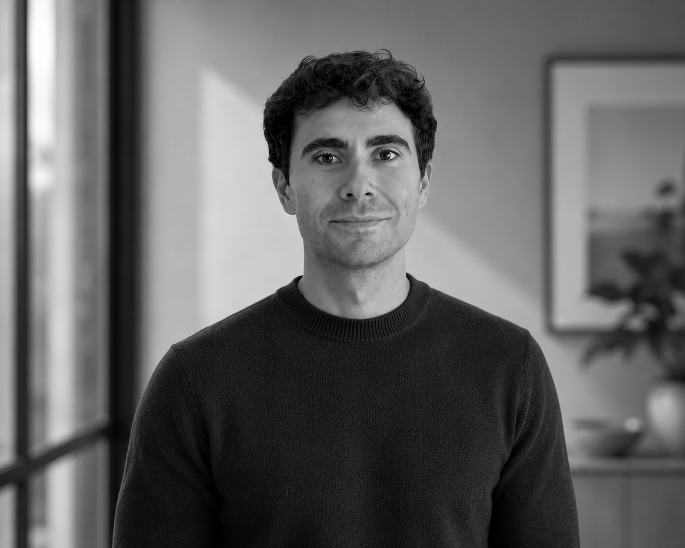 Black and white portrait of a young man with wavy hair, wearing a dark sweater, standing indoors next to a window with blurred background of a room and plant.