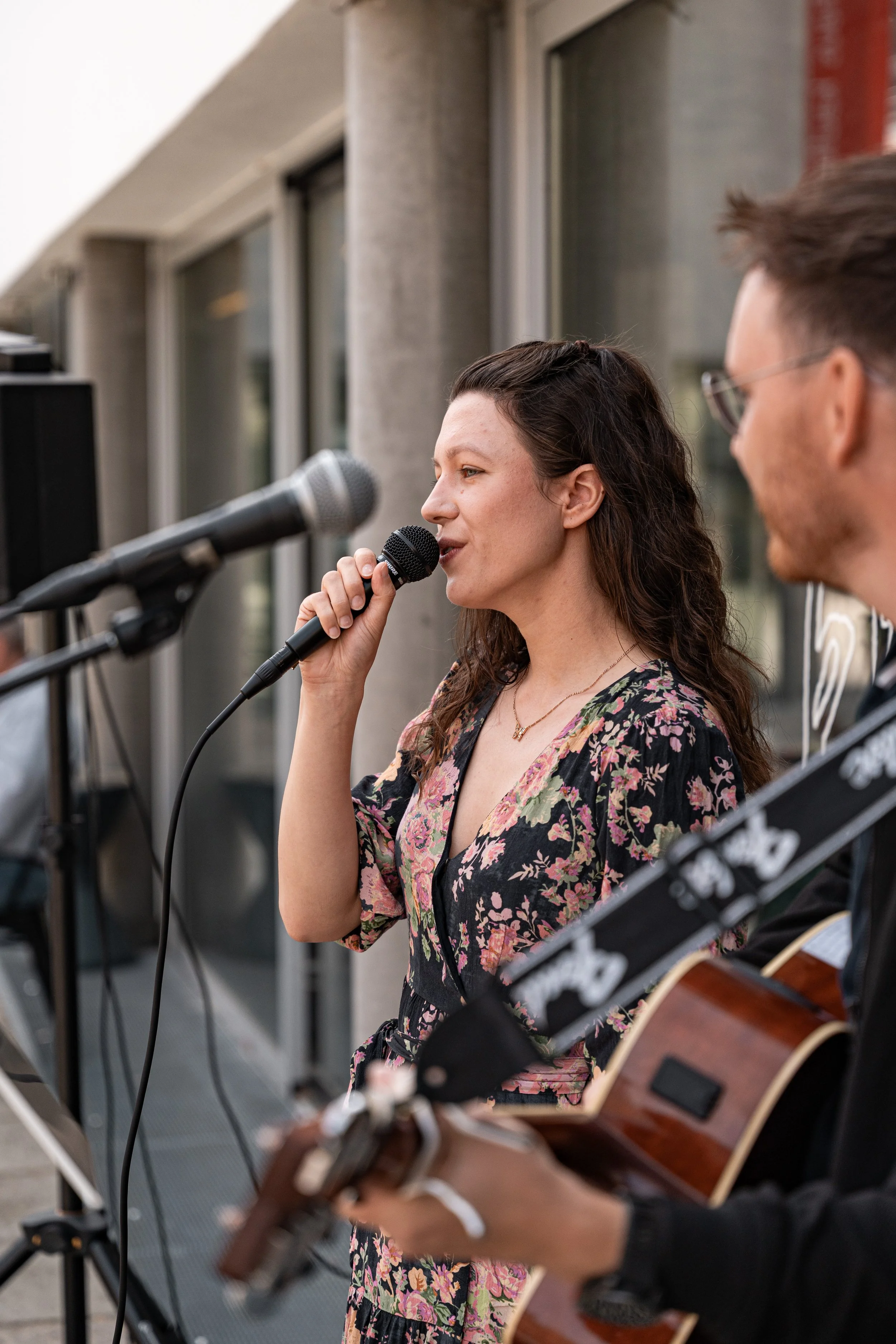 Sängerin mit Mikrofon in der Hand, die in das Mikrofon singt. Im Vordergrund der Gitarrist, verschwommen, der zur Sängerin blickt