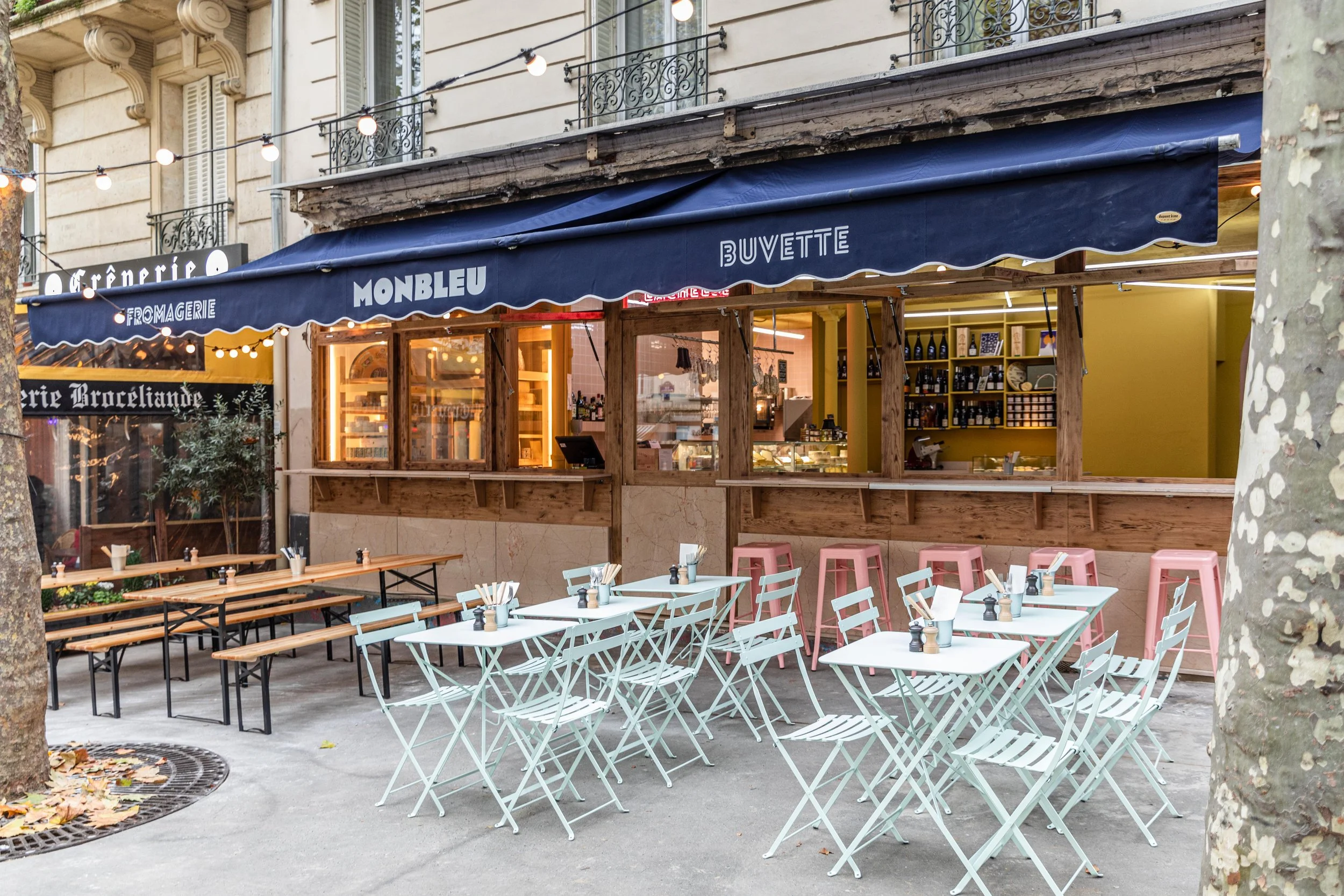 Restaurant de plein air avec des tables blanches et des bancs en bois, des chaises roses et des tables en bois, devant un bâtiment historique avec une terrasse couverte portant une bannière bleue "Monbleu Buvette".