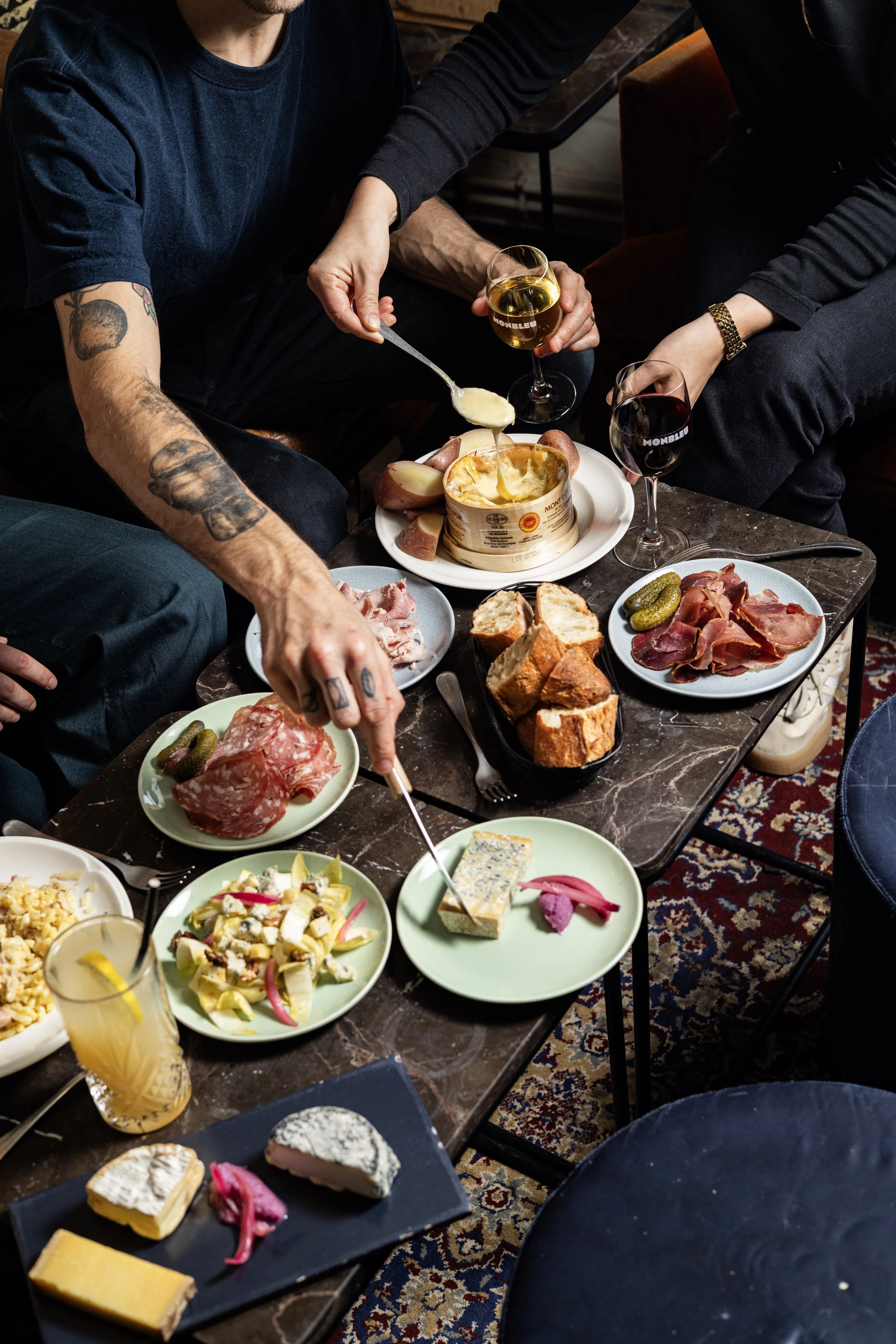 Groupe de personnes partageant un repas avec divers plats, fromages, charcuterie, pain, salades, fromages et boissons sur une table en marbre.