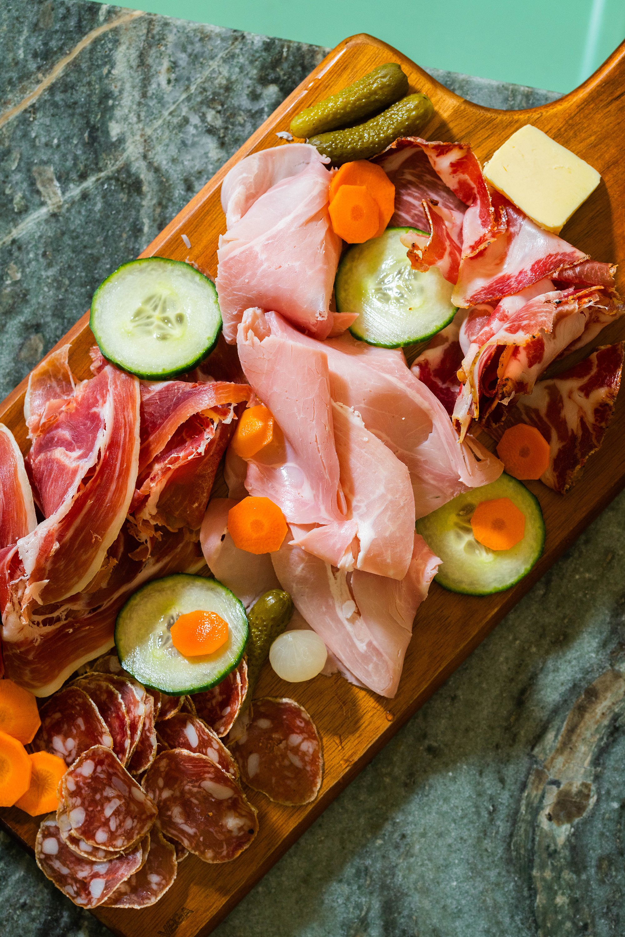 Gros plan sur une planche de charcuterie, avec des carottes en plus, des concombres, des cornichons et du beurre