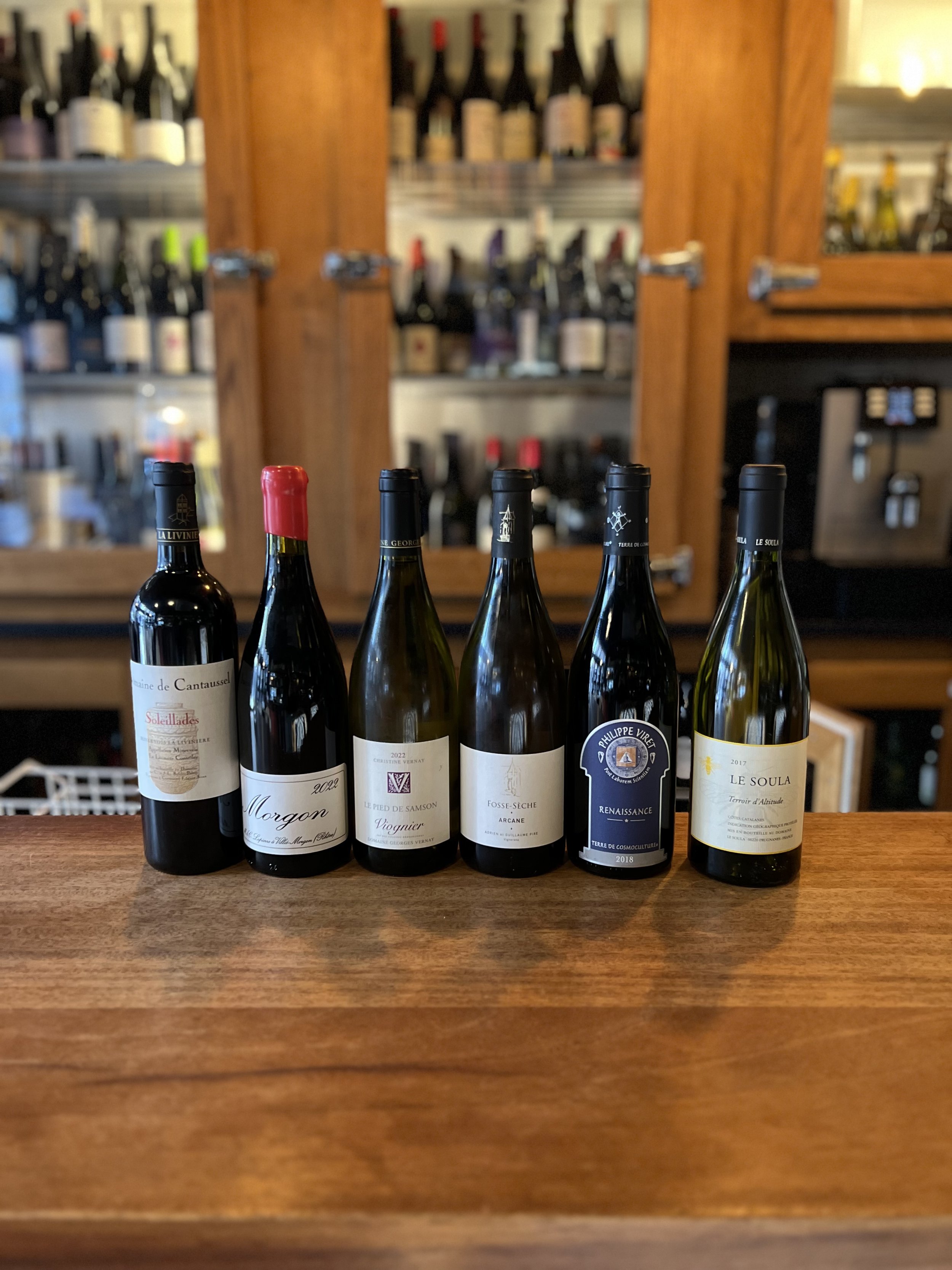 6 bouteilles de vin alignés sur une table. En arrière plan il y a plusieurs vitrines avec des bouteilles de vin rangés