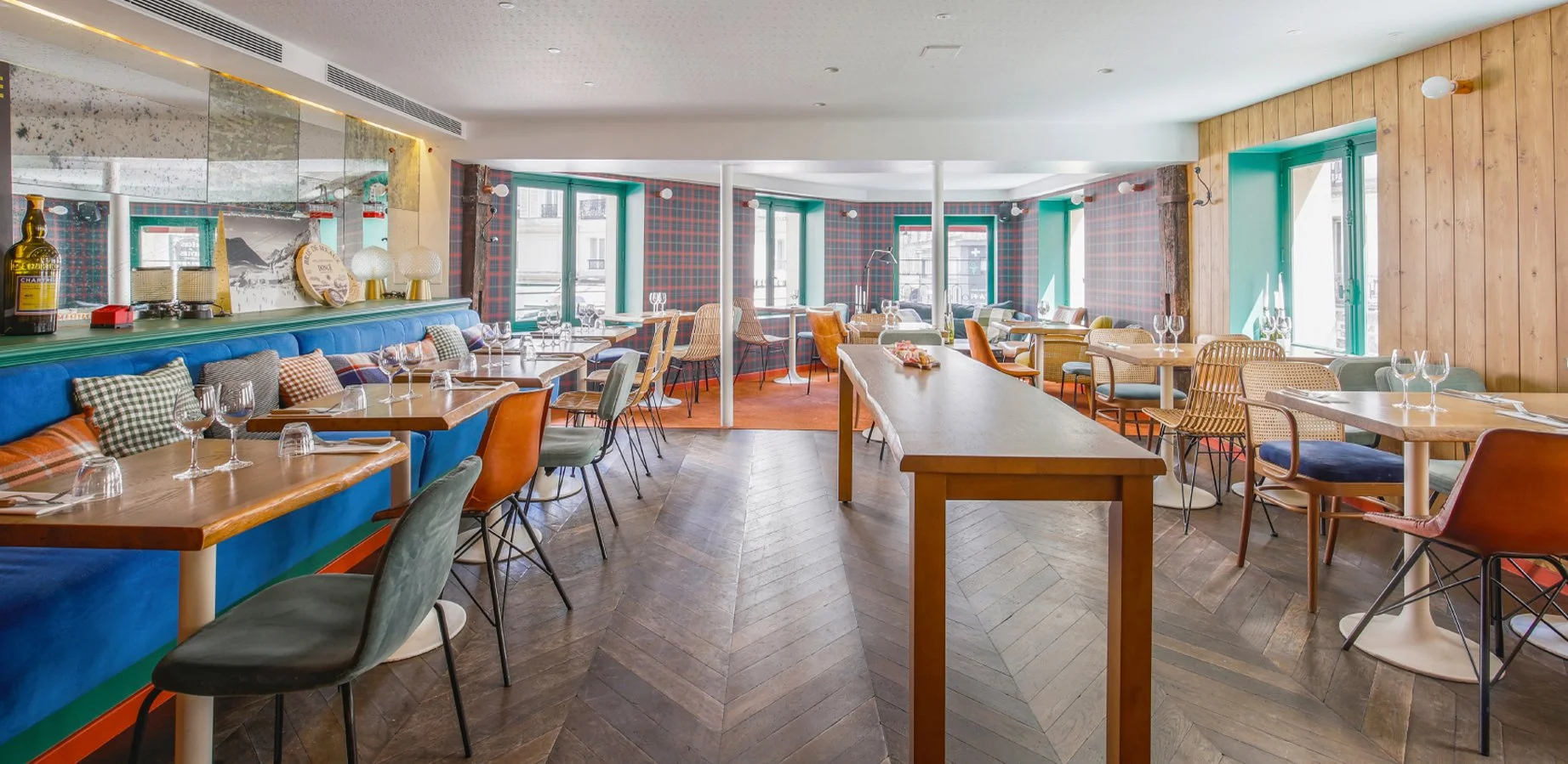 Salle à manger élégante avec tables en bois, assiettes et verres, chaises variées, murs en bois et carreaux, grandes fenêtres laissant entrer la lumière naturelle, décor coloré et confortable.