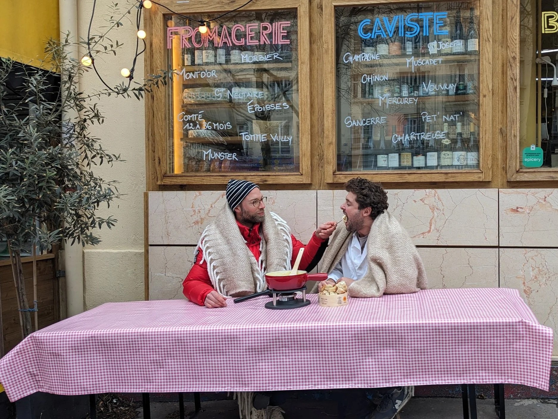 Deux hommes mangent une fondue, ils ont des couvertures sur eux, l'un met dans la bouche de l'autre une fourchette rempli de fondue de fromage