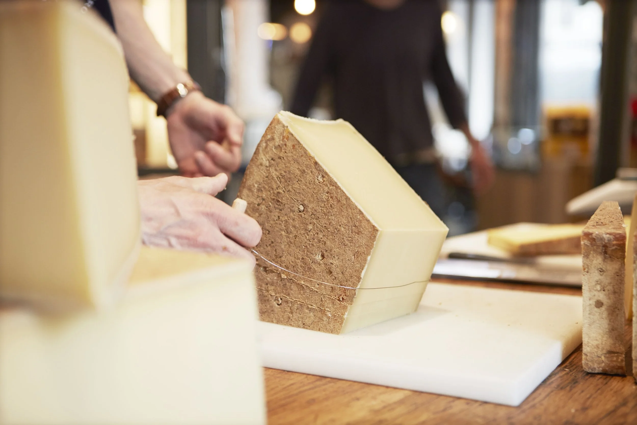 Gros gros plan sur un fromage avec une tranche coupée, posé sur un plan de travail en bois. En arrière-plan, deux personnes manipulant le fromage, floues.