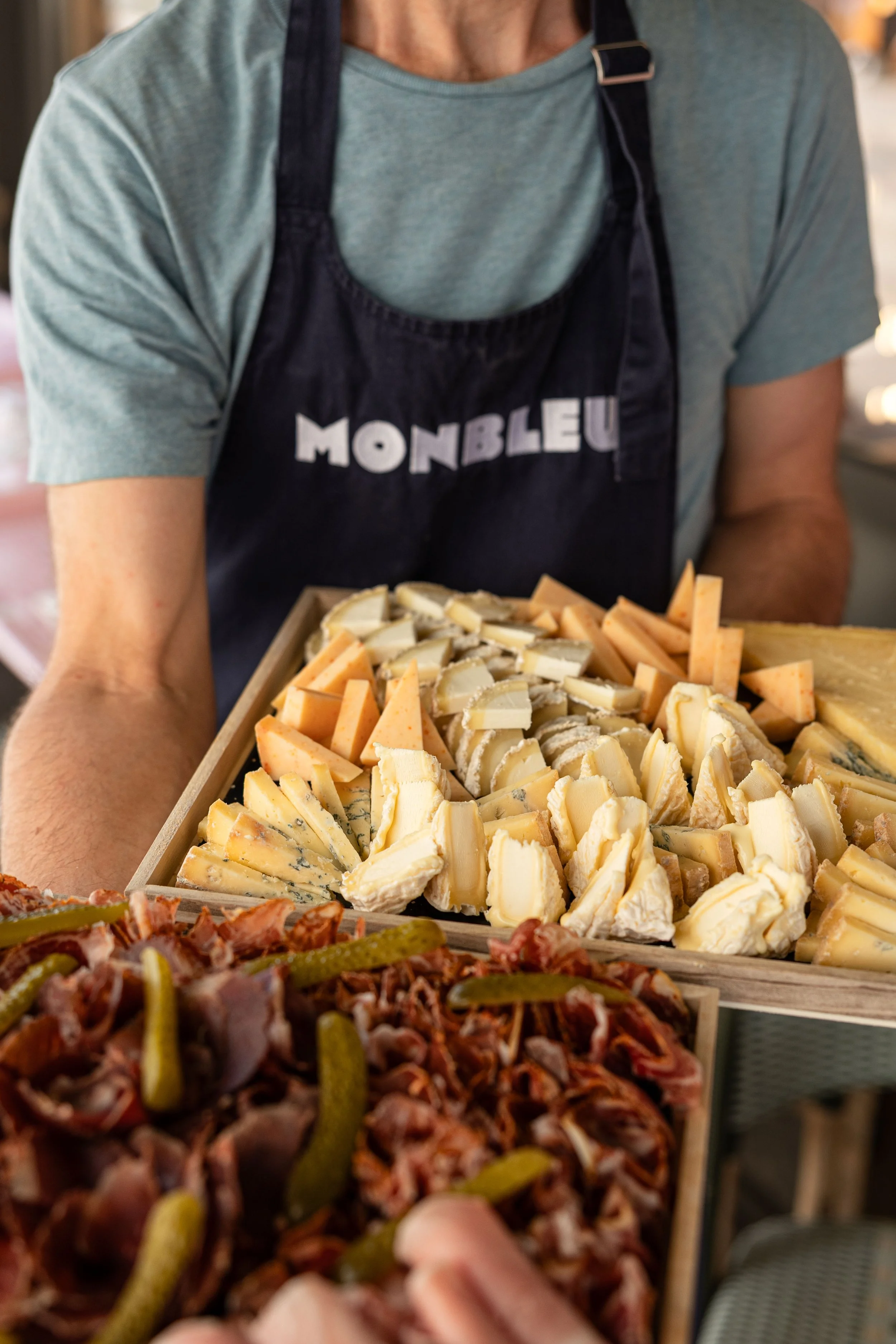Une personne portant un t-shirt vert et un tablier noir avec écrit "monbleu" propose un plateau de fromages variés et un plateau de charcuteries variés, avec des cornichons