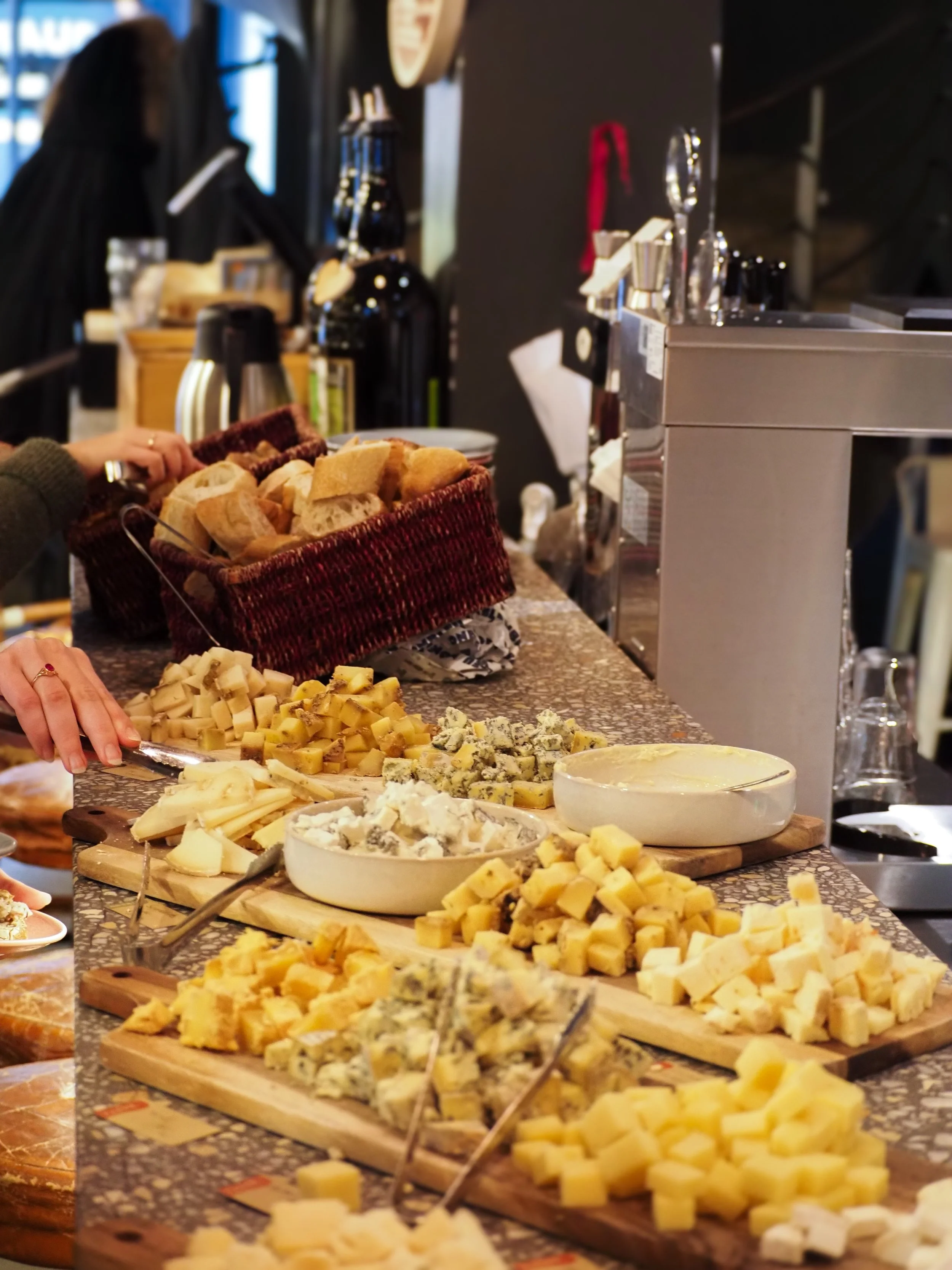 Le brunch du dimanche avec pleins de fromage à volonté posé sur des planches, des personnes se servent, il y a plusieurs corbeille à pain. 