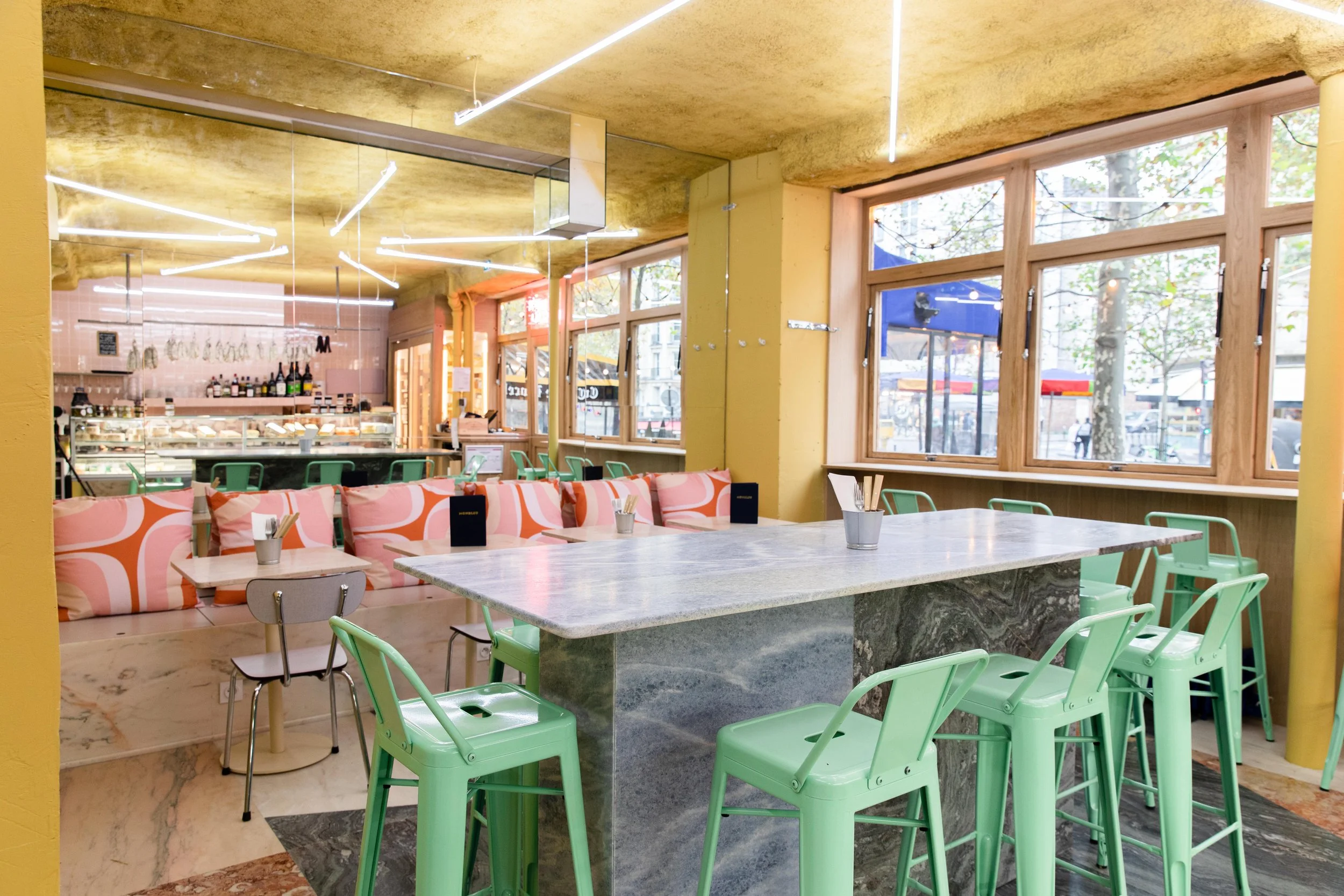 intérieur d'un restaurant très lumineux, avec des tables clair et des chaises vertes, les meubles sont en marbres et il y a des coussins roses. Il y a un grand miroir qui nous permet de distinguer la fromagerie avec tous les produits proposés
