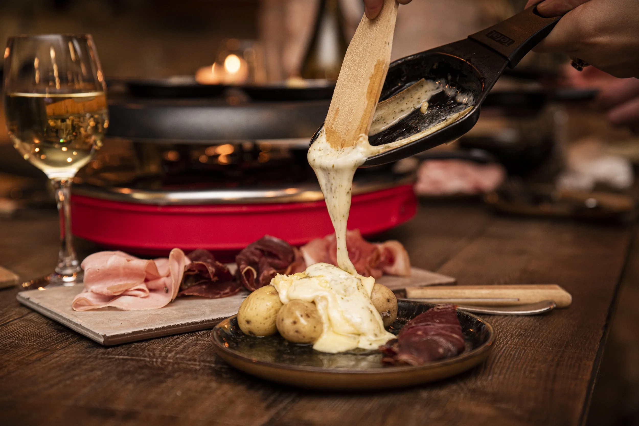 Un verre de vin blanc à gauche, une petite planche de charcuterie, une assiette avec des pommes de terre et de la charcuterie. Par dessus l'assiette une personne verse de la raclette fondue sur les pommes de terre. En arrière plan il y a l'appareil à