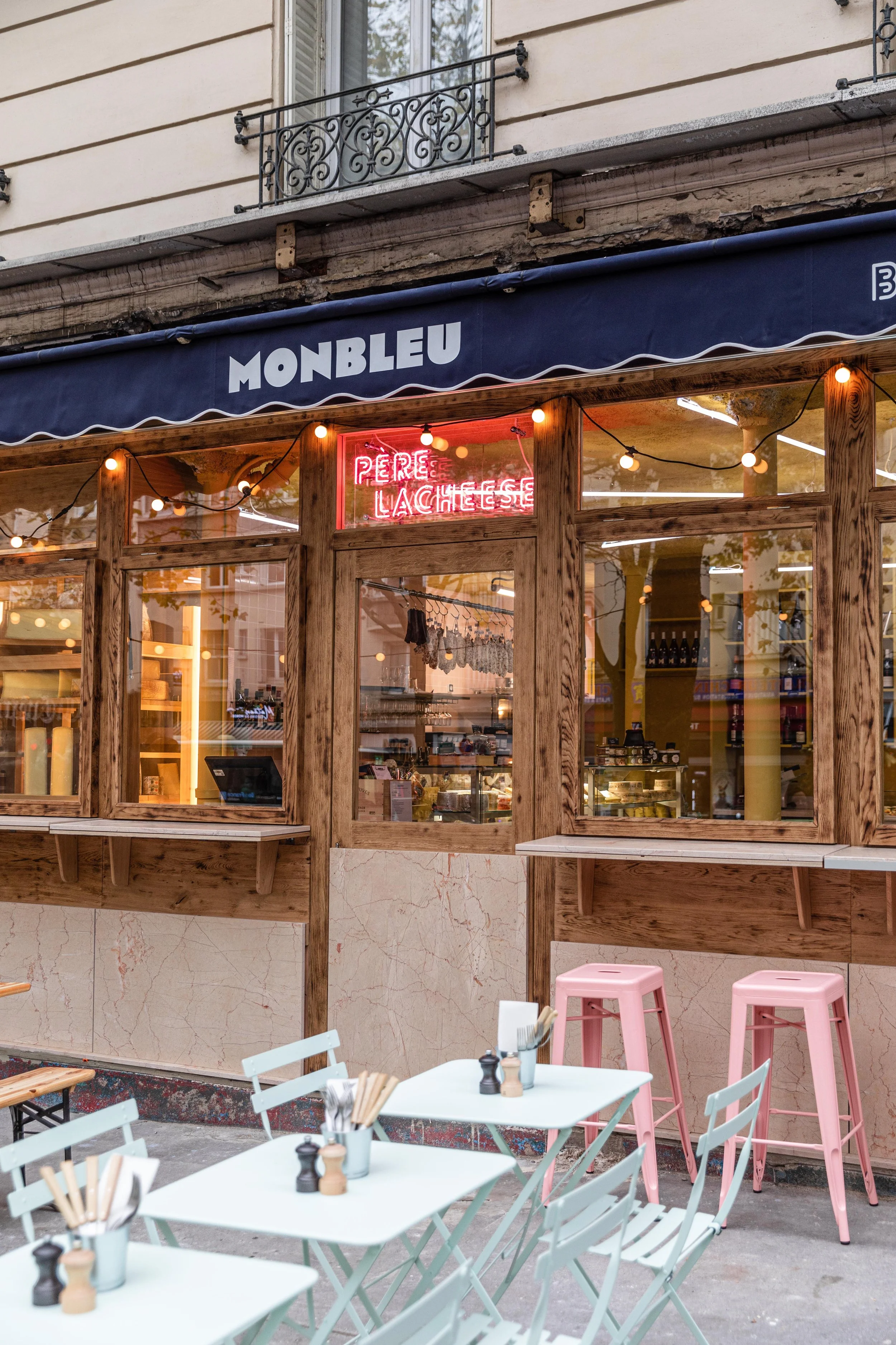 Sur la devanture il y a le nom du restaurant "monbleu père lacheese" , on voit la terrasse on est a l'extérieur du restaurant, les tables et chaise sont roses et bleus, ton pastel