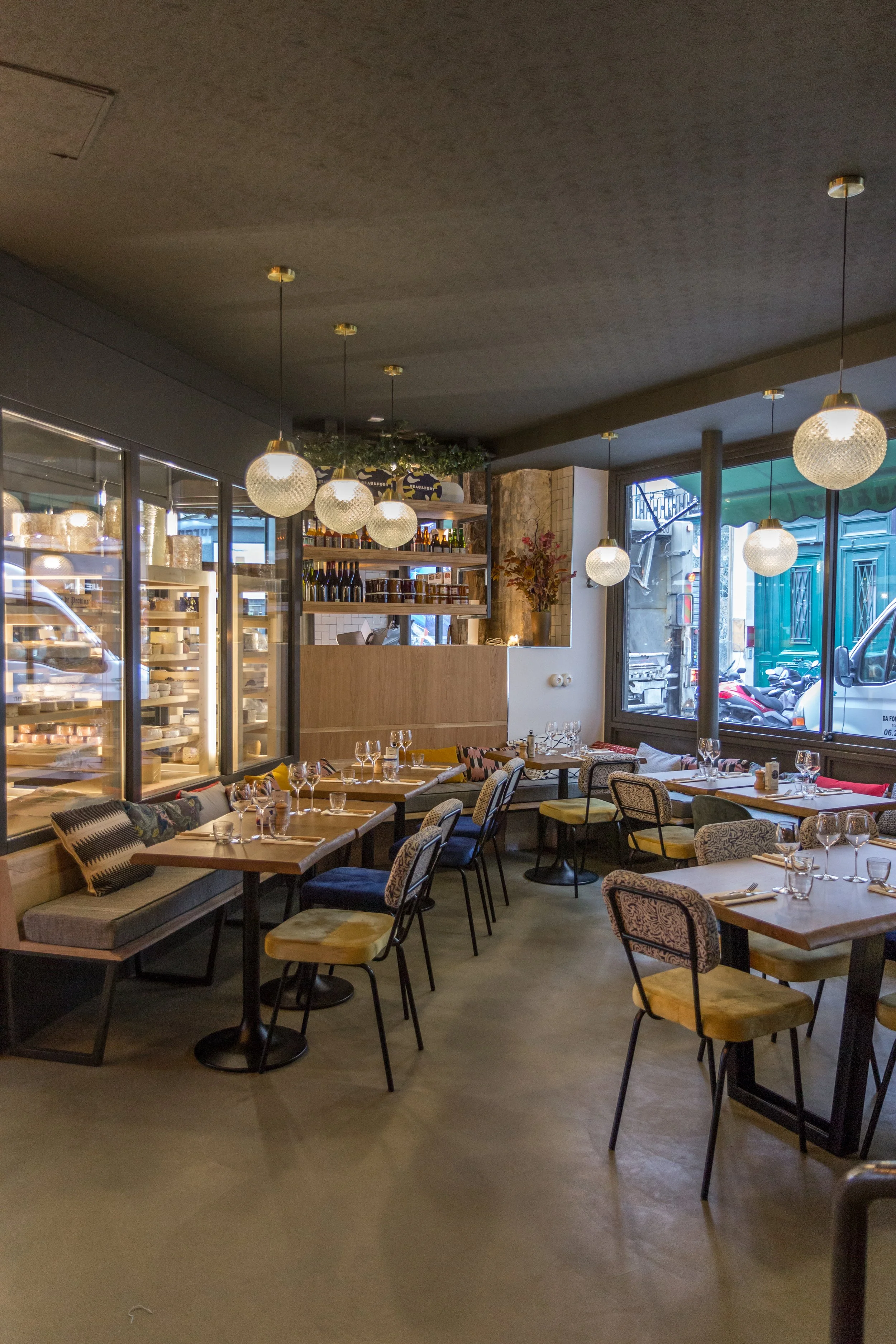 Intérieur d'un restaurant avec des tables, des chaises, des luminaires suspendus et une vitrine réfrigérée contenant des fromages
