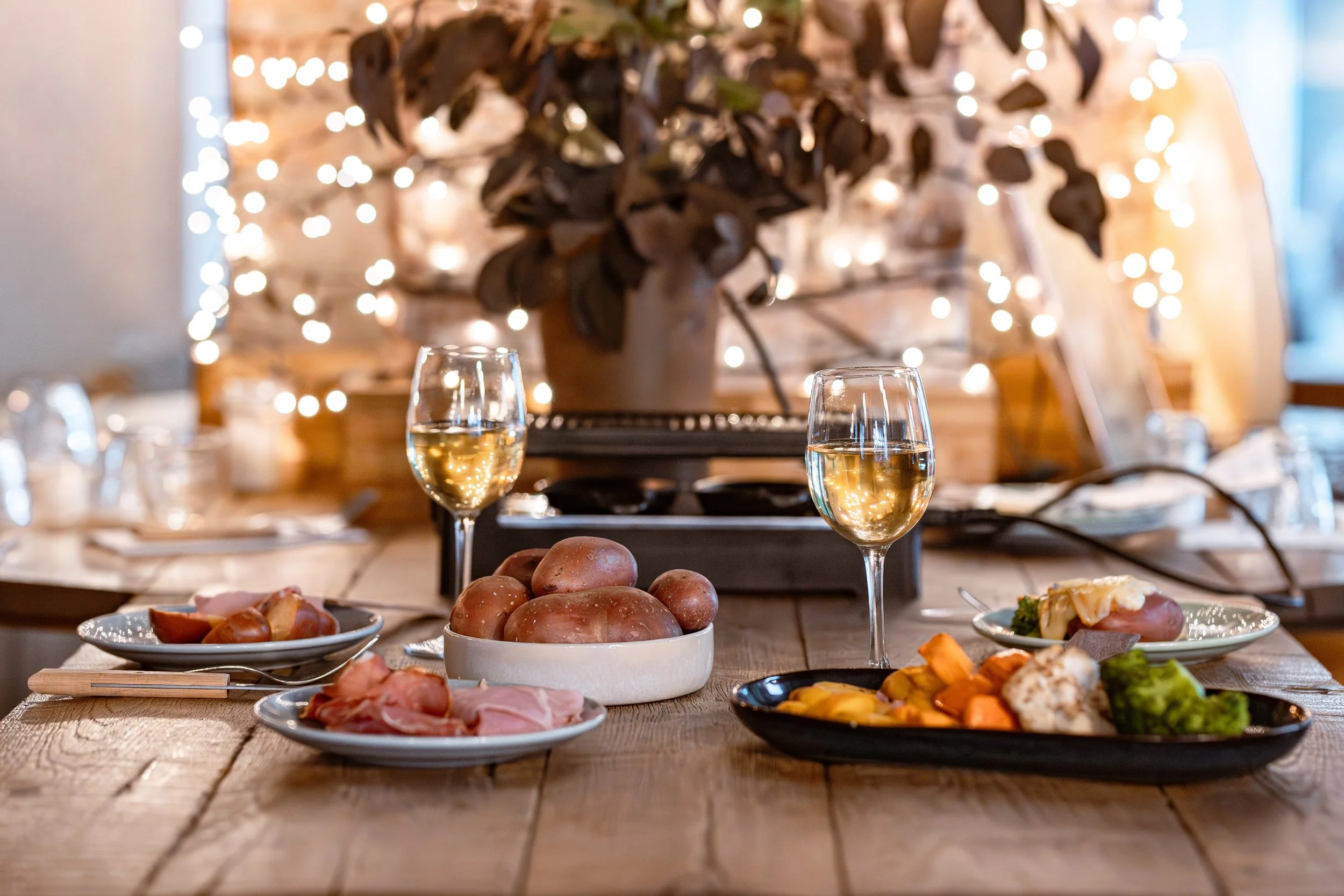 Une table dressée avec des pommes de terres, de la charcuterie, de verres à vin pleins, un appareil à raclette au fond , des guirlandes, et une plante an arrière plan, période de fête