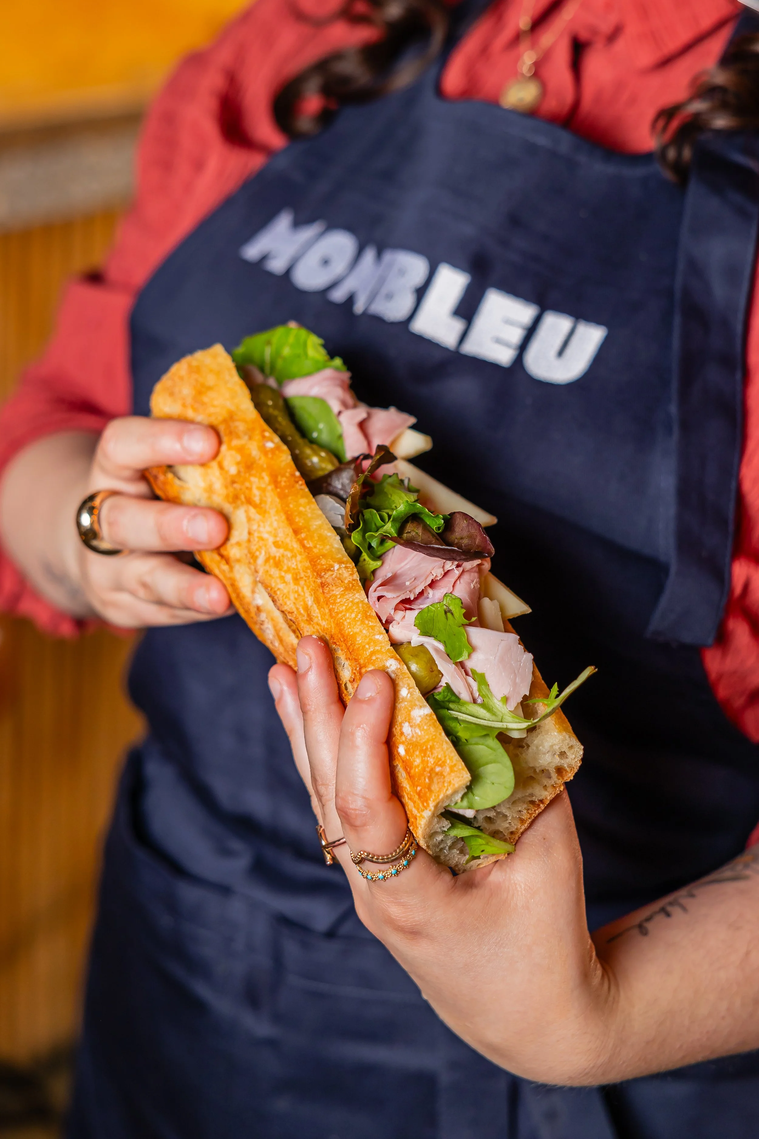 Un femme tient un sandwich bien garnie de cornichons, de jambon et de salade
