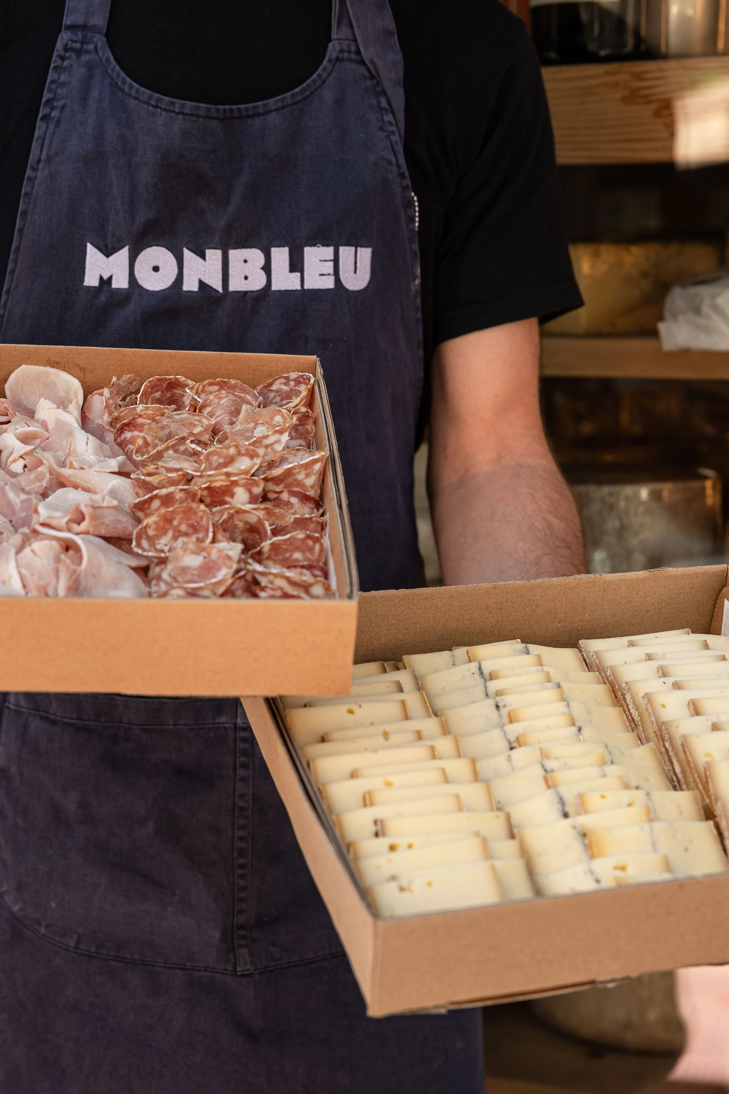 un monsieur munit d'un tablier "monbleu" tient deux plateaux, un rempli de charcuteries et l'autre rempli de fromage à raclette