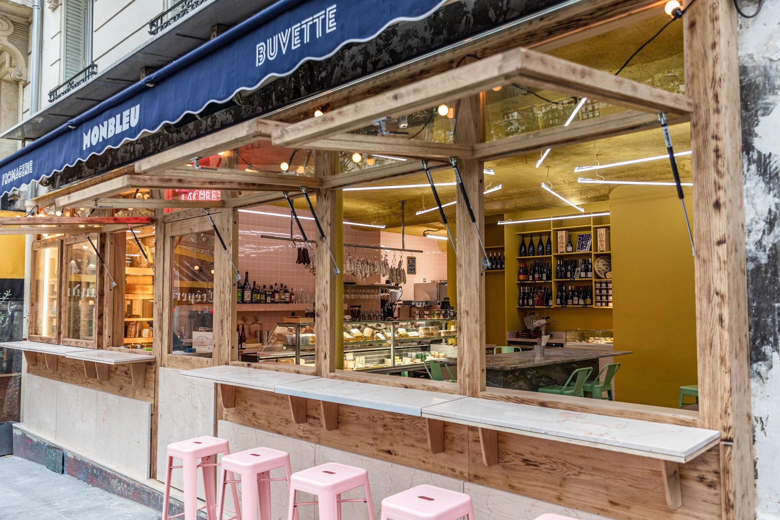Une petite boutique de fromages avec un comptoir de vente, dans un style moderne avec un en-cas à l'extérieur, des tables et des chaises en plastique rose, un mur jaune avec des bouteilles de vin en étagères.