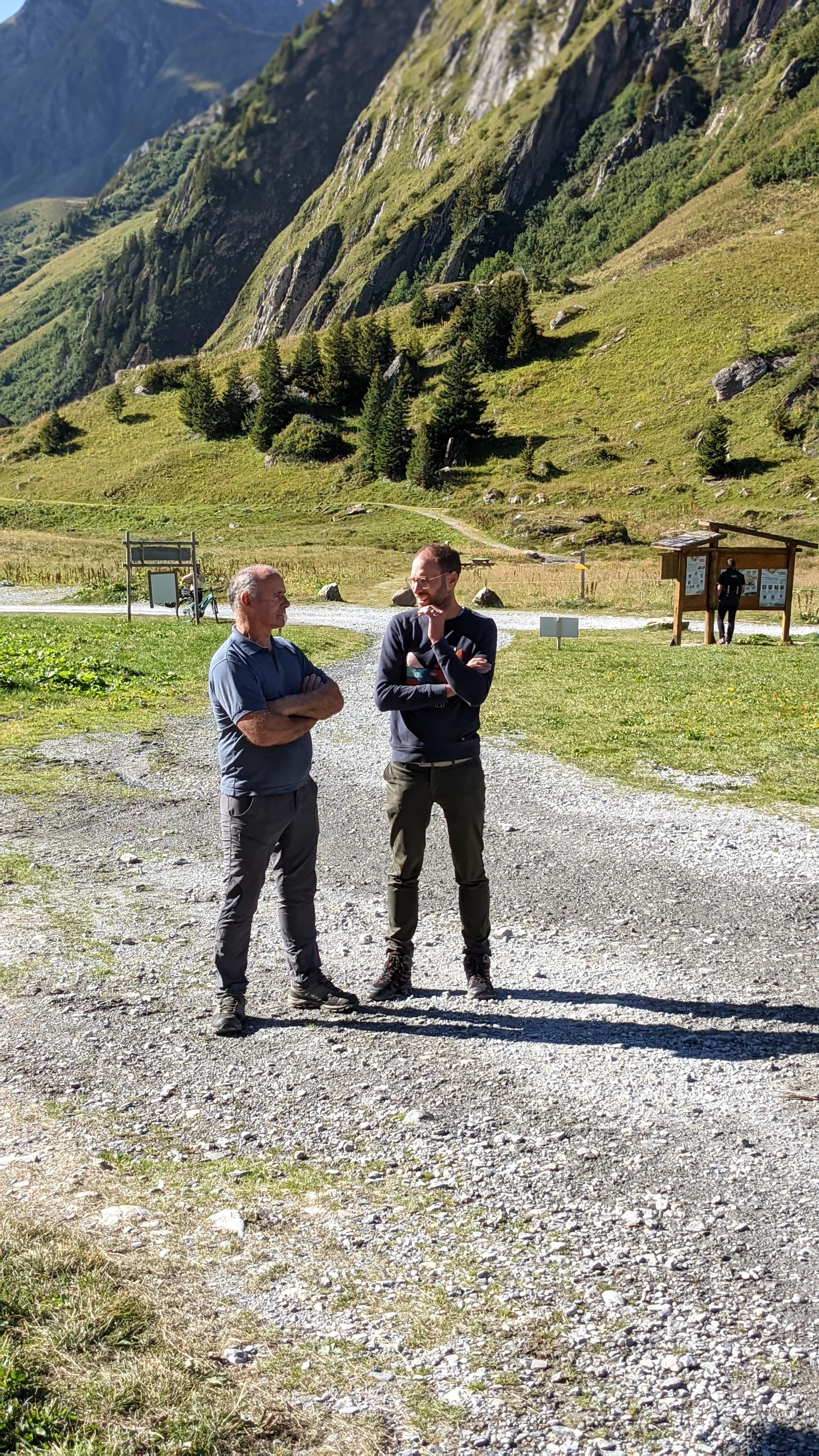 Deux hommes discutent dans un sentier de montagne entouré de verdure et de rochers, avec des montagnes en arrière-plan, par une journée ensoleillée.